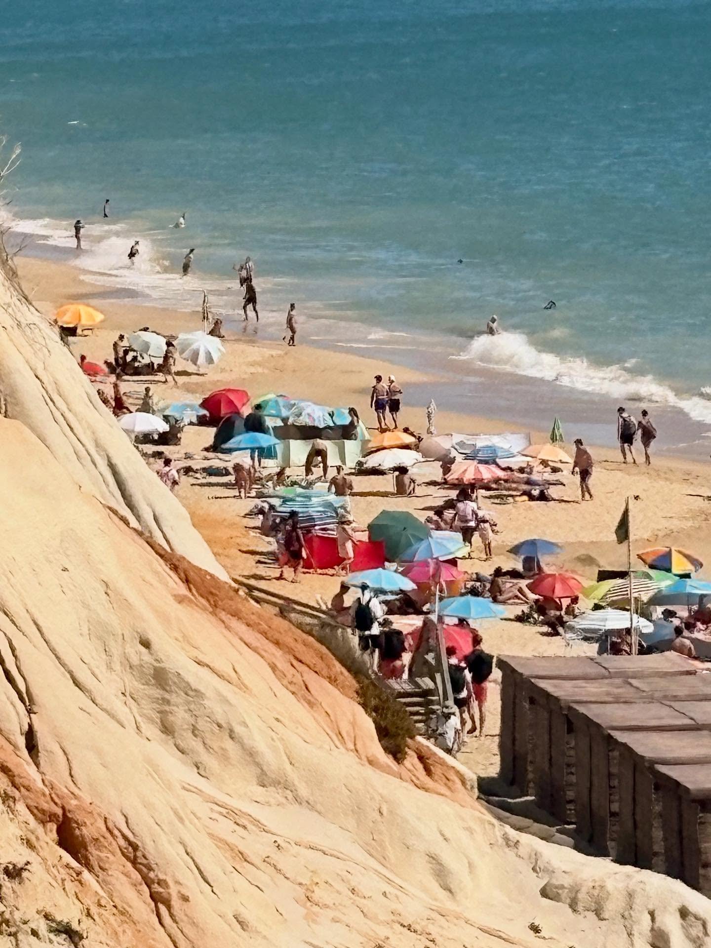 Beach with many colorful umbrellas, people walking and relaxing on the sand, and shoreline with waves, viewed from a cliffside.