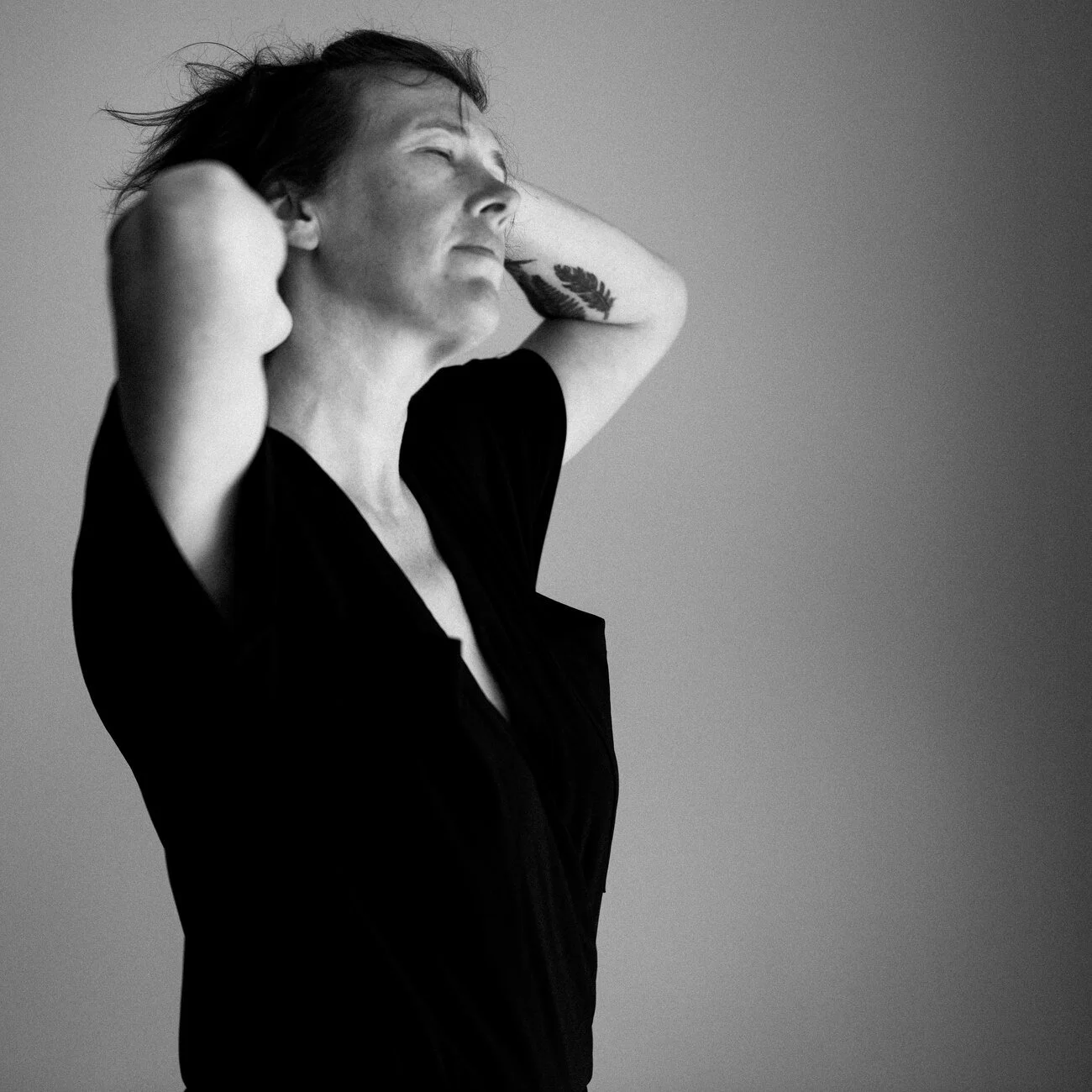 A woman with short hair and a tattoo on her arm, wearing a black shirt, standing against a plain background with her eyes closed and hands behind her head.