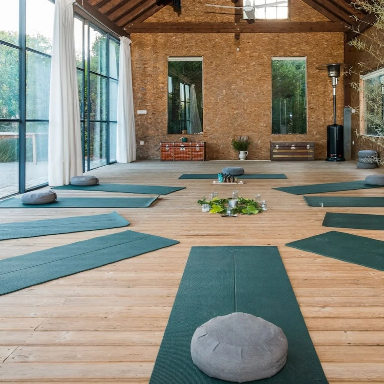 A spacious indoor yoga studio with wooden floors and large glass windows, arranged with green yoga mats and round cushions, and decorated with plants and candles.