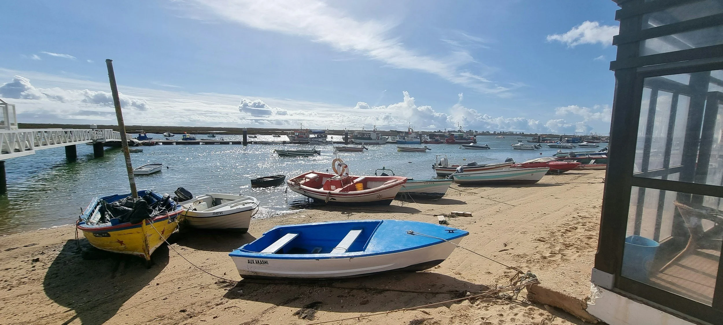 A sunny day at a marina with small boats on the sandy shore and in the water, clouds in the sky, a white pier on the left, and a glass structure on the right.