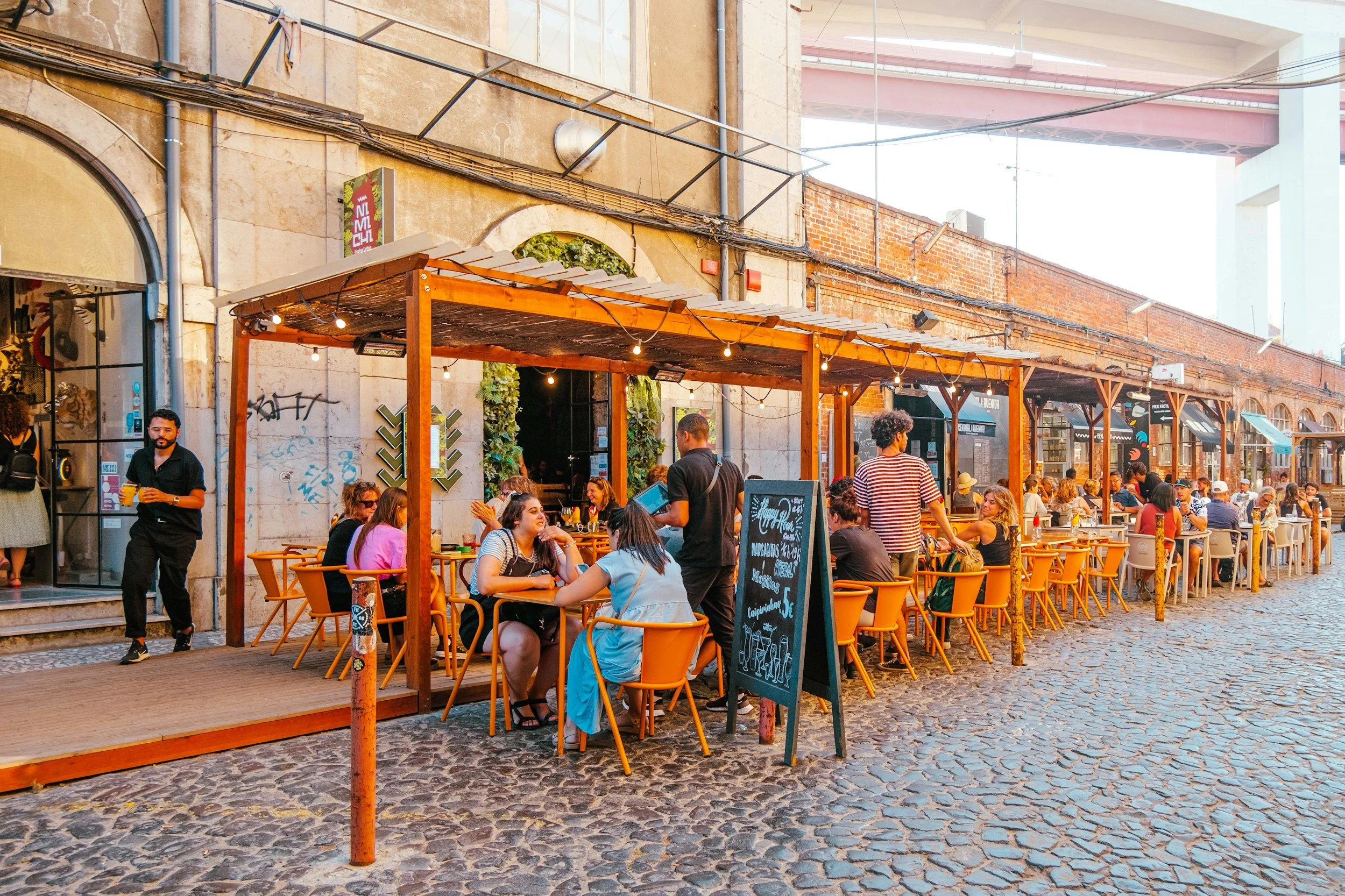 People dining at an outdoor patio of a restaurant under a wooden pergola in an urban setting during daytime.