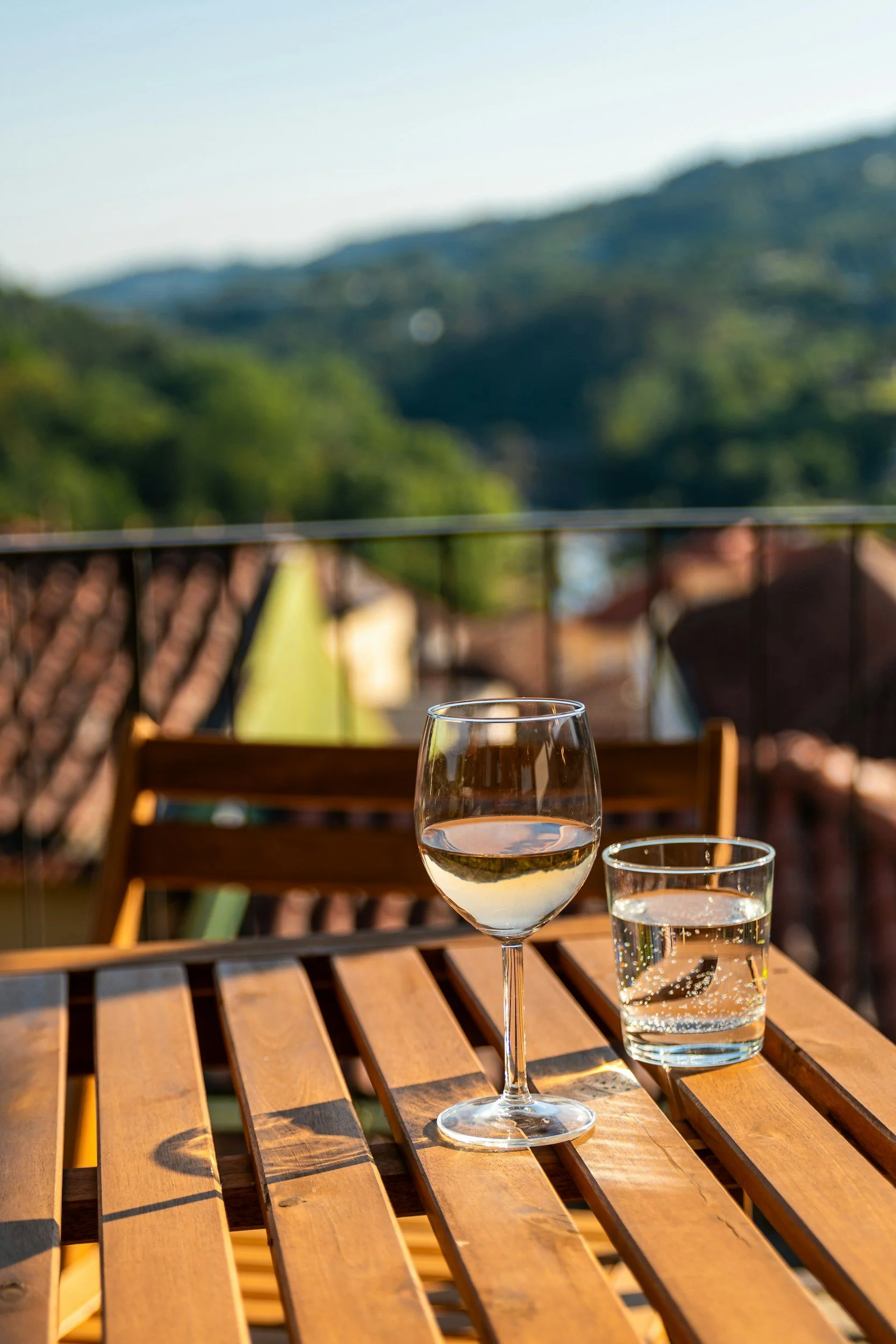 A table with a glass of white wine and a glass of water on a wooden outdoor table, with a blurred background of a balcony railing, rooftops, and mountain range.