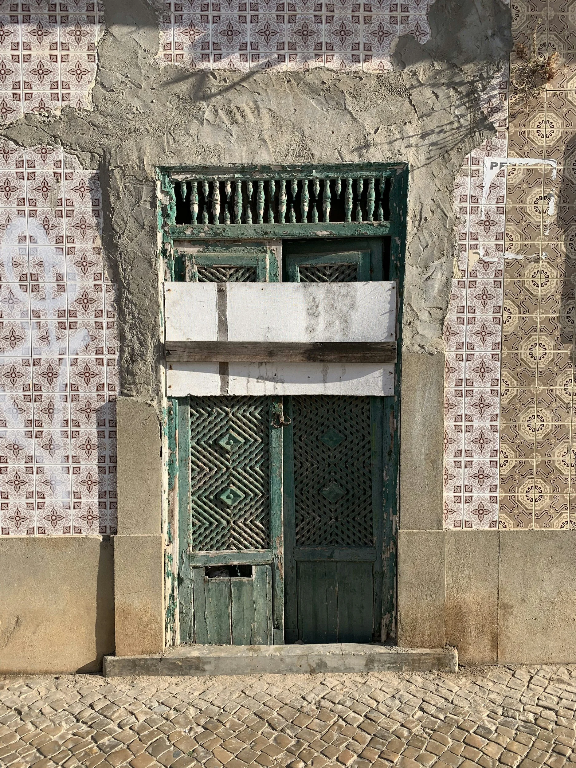 An old, weathered wooden and metal door with peeling green paint, set in a concrete and tiled wall. The wall features decorative patterned tiles on both sides, with the left side having red and white geometric design tiles, and the right side featuri