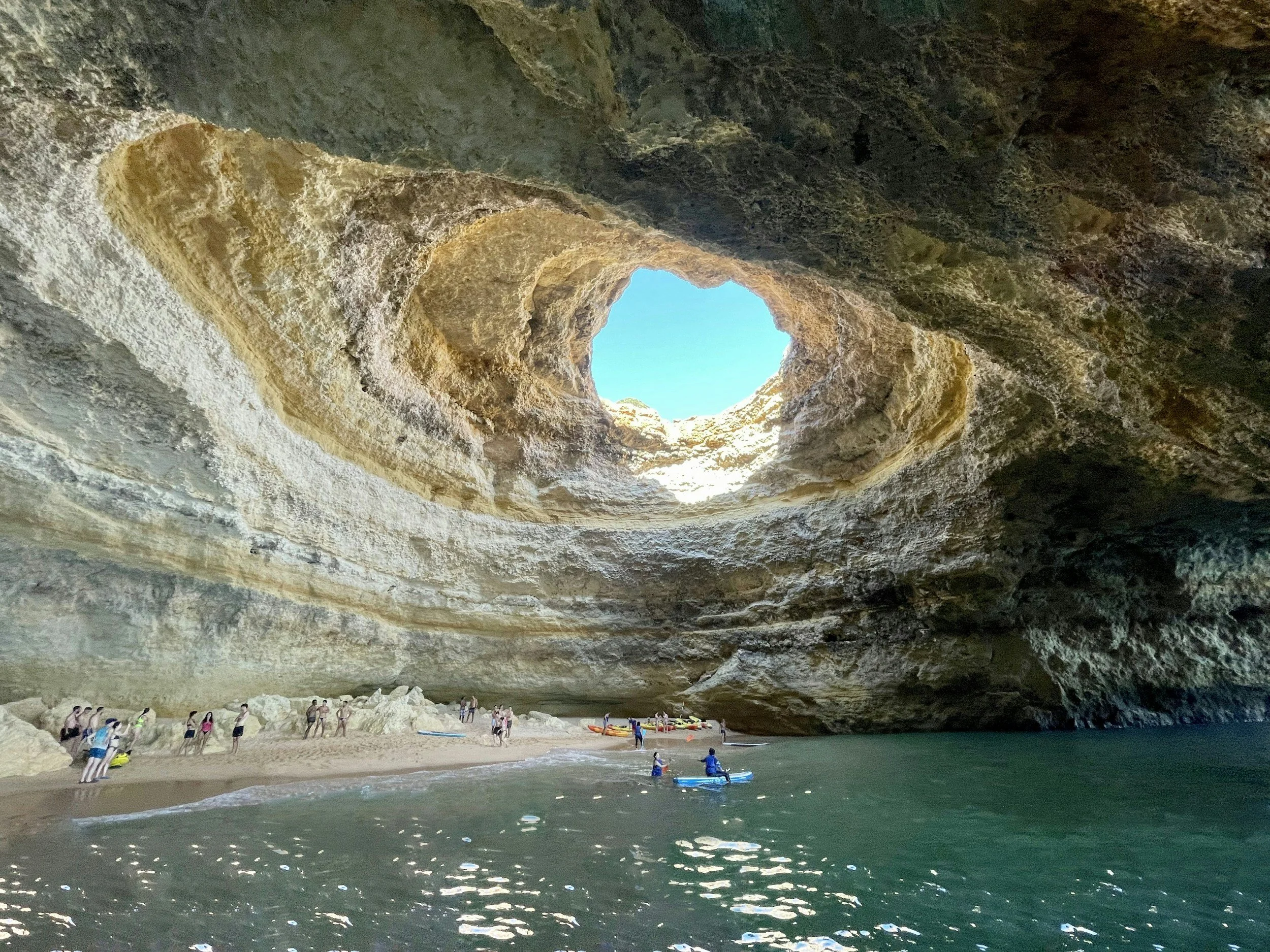 A large cave with a sandy beach and people kayaking and exploring near the water, with the cave's opening showing a bright blue sky.