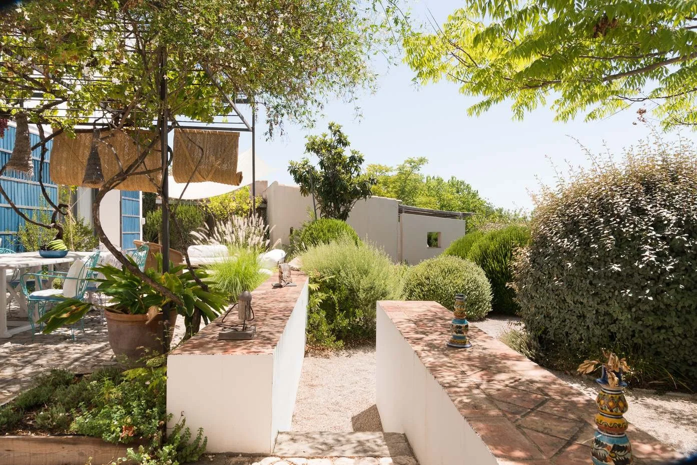 A bright outdoor garden with lush green bushes and trees, white and blue furniture, and decorative pottery on tiled and white surfaces, under a clear sky.