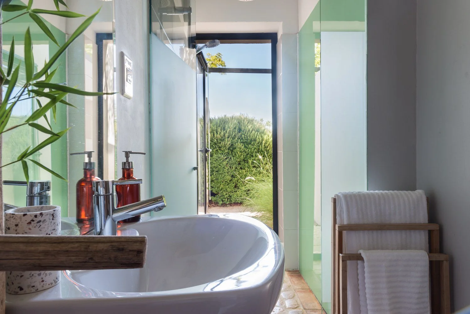 A modern bathroom with a white sink, amber soap dispensers, a toothbrush holder, and a small plant. Outside the window, green bushes are visible, and part of a garden or outdoor area. Towels are on a wooden rack next to the sink.