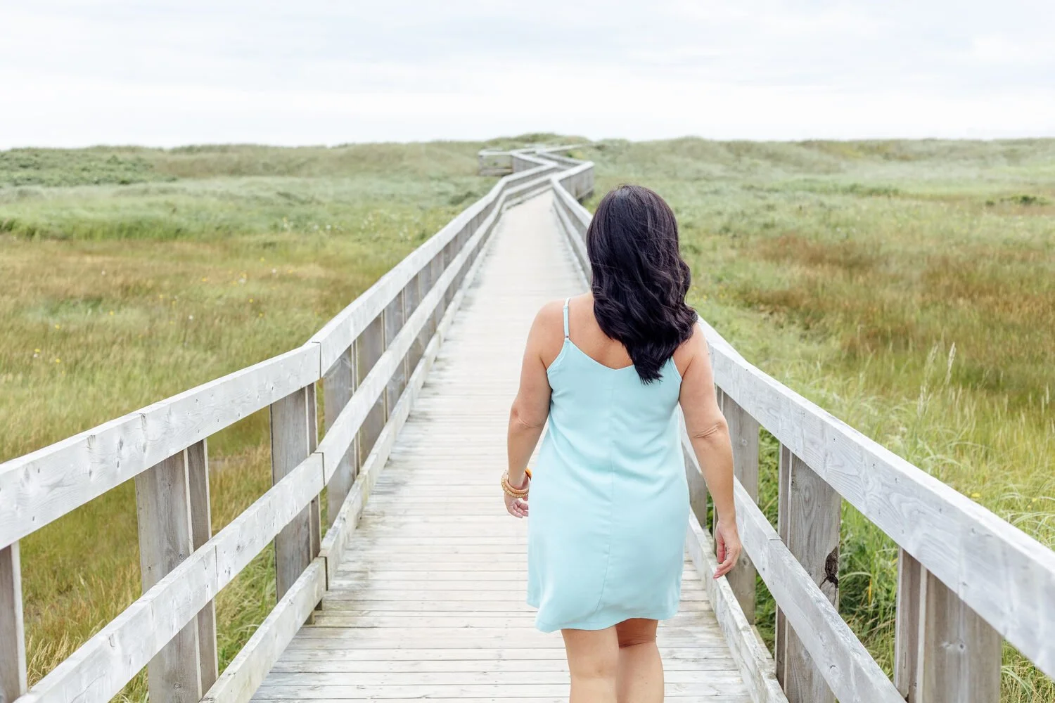 Charmaine Renee, rest coach and breathwork practitioner, walks on a wooden boardwalk through a green grassy field towards the horizon on a cloudy day.
