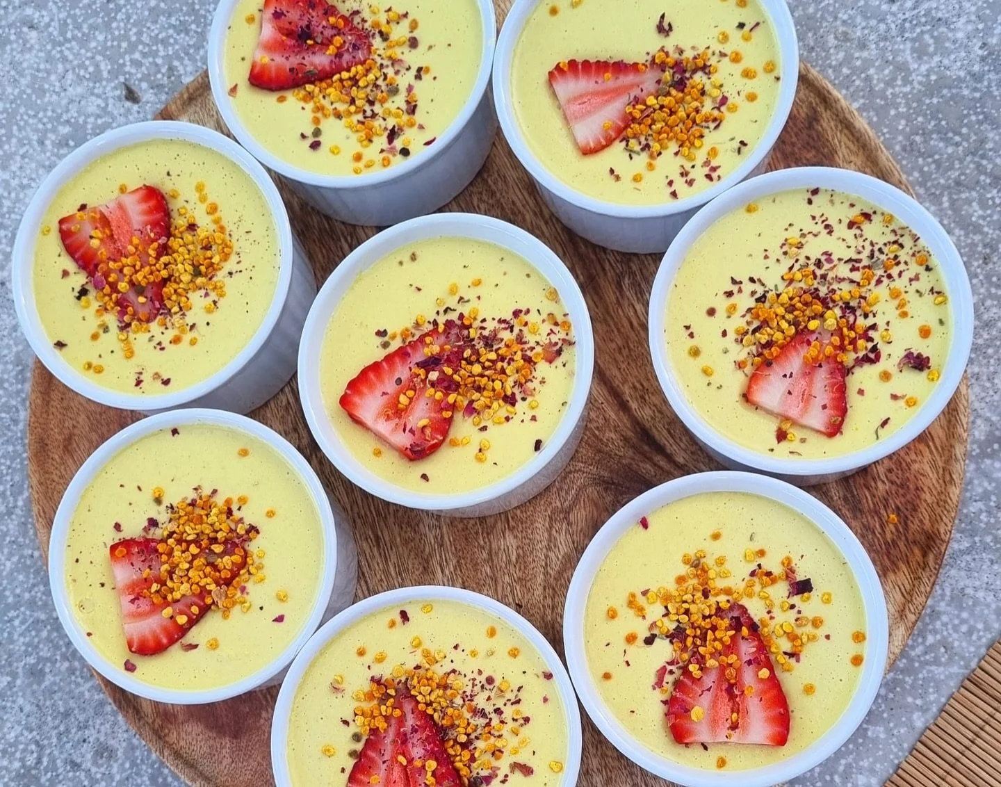 Eight cups of yellow dessert topped with sliced strawberries and orange and purple sprinkles, arranged on a round wooden tray.