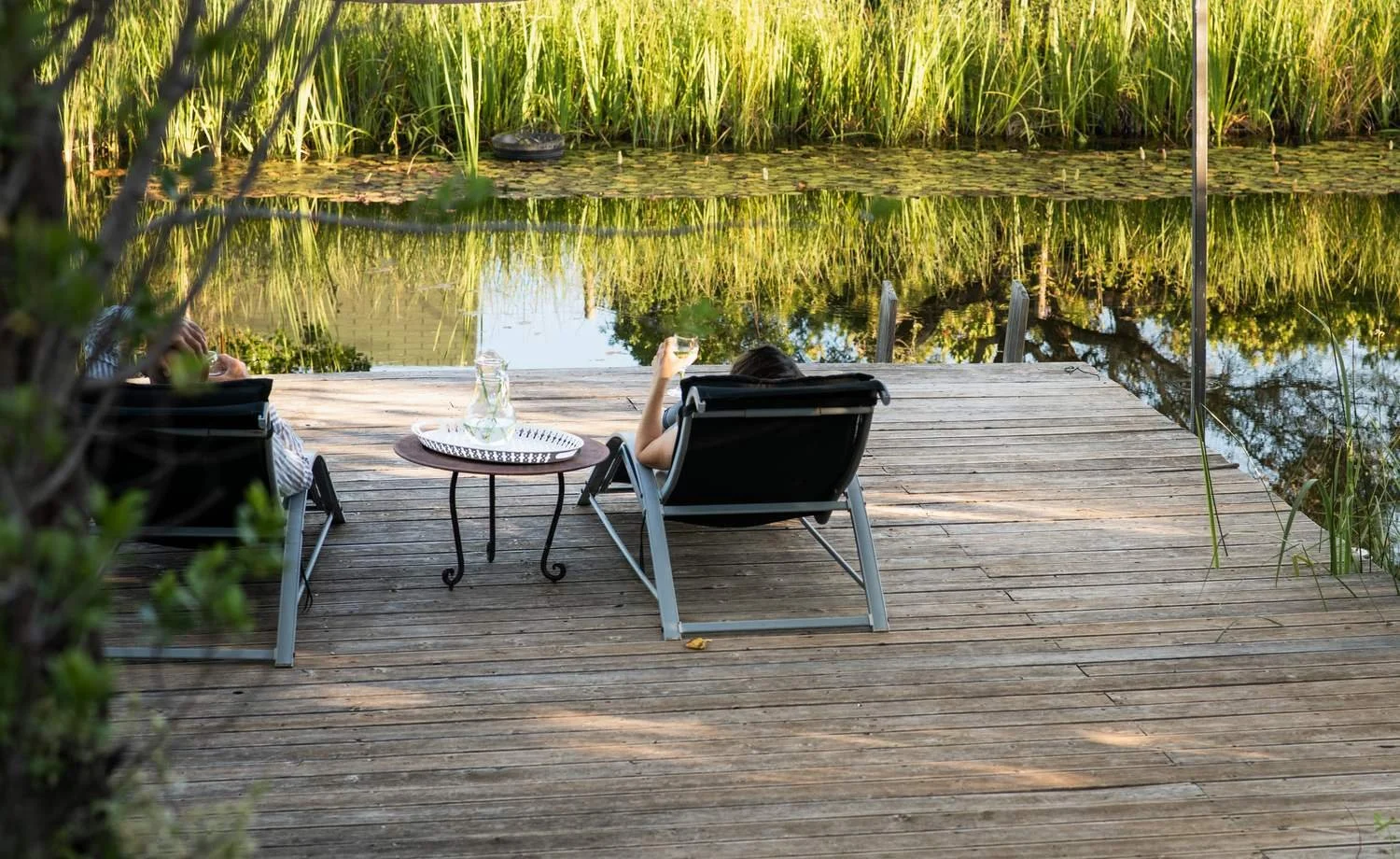 Two people relaxing on lounge chairs on a wooden dock beside a pond, with one person holding a glass and a tray with a water pitcher nearby. The pond is surrounded by tall green grass, and the scene is peaceful and scenic.
