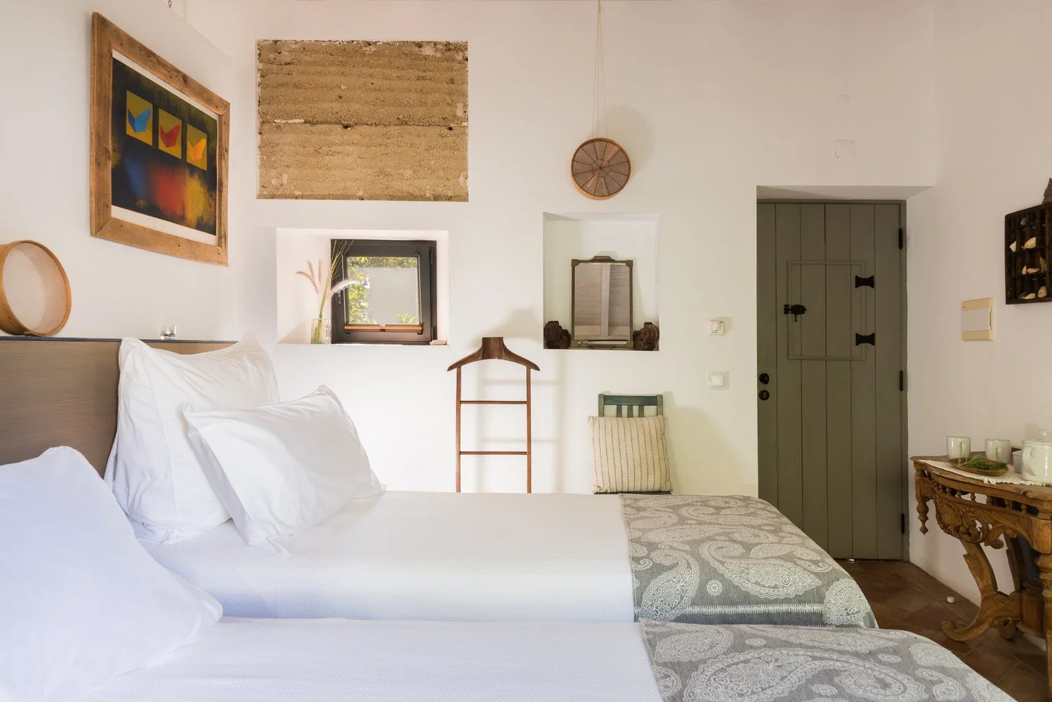 A cozy bedroom with two beds, white linens, decorative pillows, and a textured curtain on one bed, against a white wall with artwork and decorations, small window with a view outside, and vintage furniture including a wooden table with cups and a mirror in a cottage-style setting.