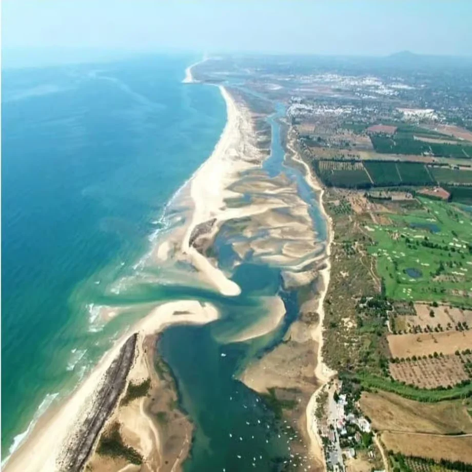 Aerial view of a coastline with sandy beaches and a river flowing into the ocean, surrounded by farmland and small buildings.