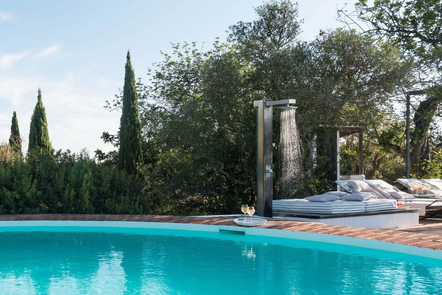 Outdoor swimming pool with lounge chairs, towels, and a shaded shower outside, surrounded by trees and greenery under a partly cloudy sky.