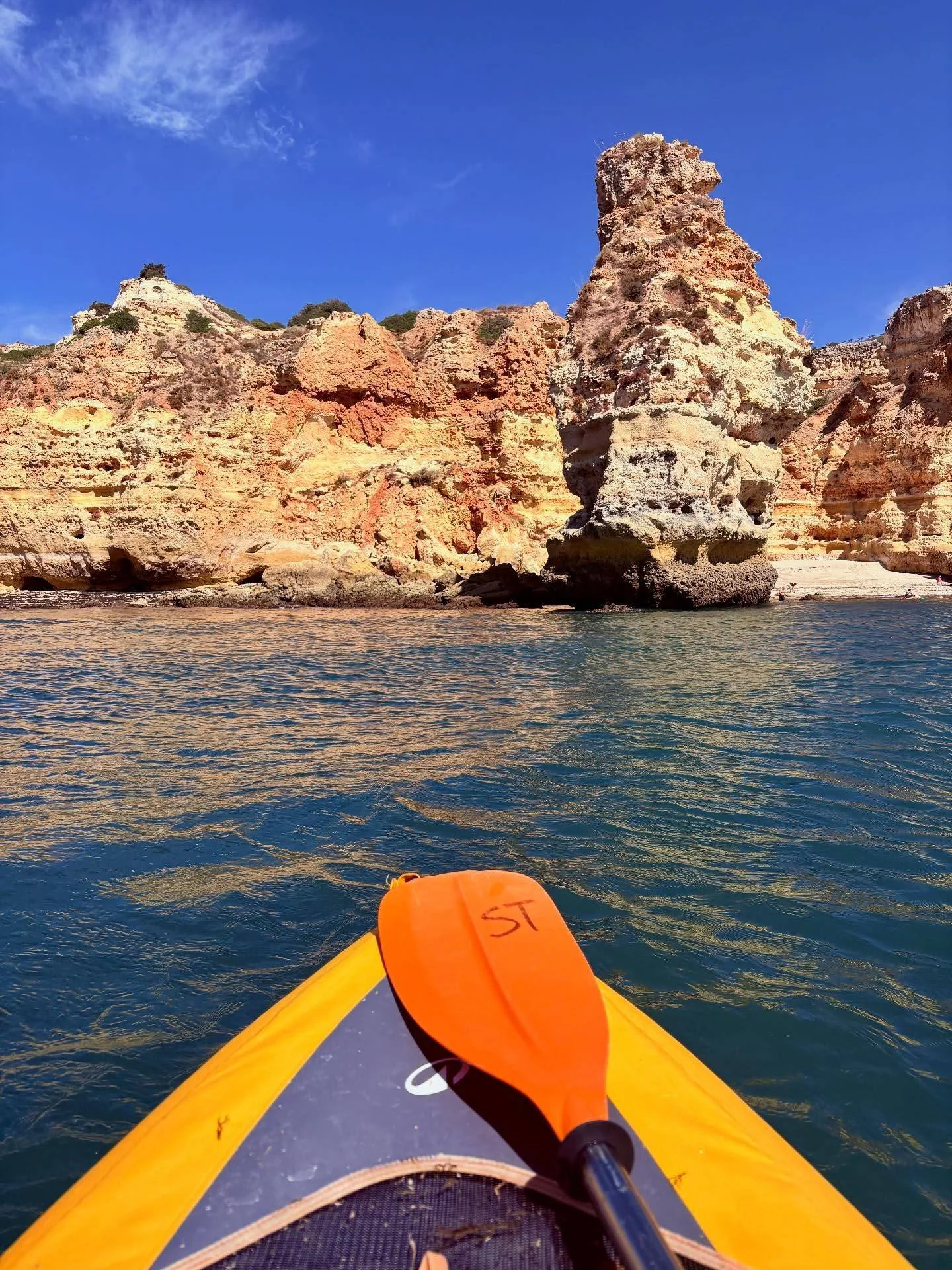 View from a kayak on blue water, facing a rocky shoreline with colorful cliffs and a large rock formation, under a clear sky.