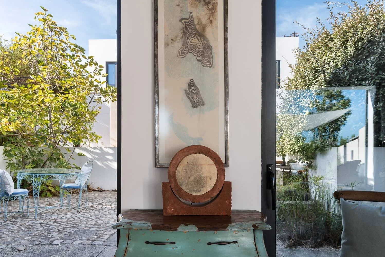 View through a glass door to an outdoor patio with trees, a white wall, and a metal table with chairs. Inside, a decorative art piece and a sculpture sit on a distressed wooden table against a white wall.