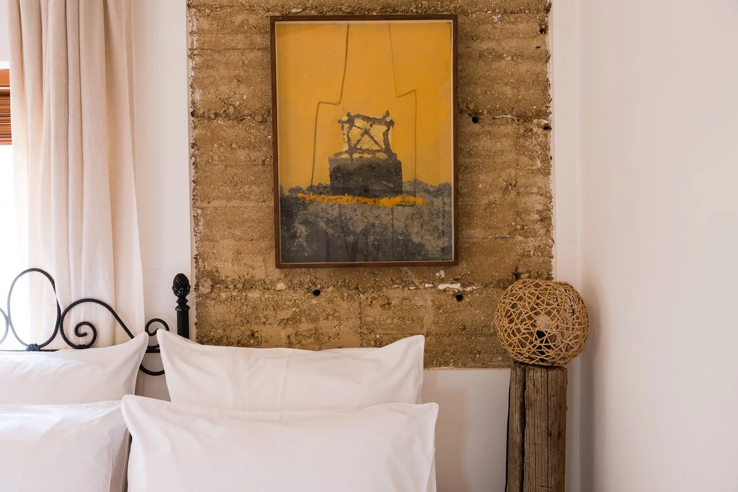 A bedroom corner featuring a bed with white pillows, a framed abstract painting on a textured brick wall, a woven spherical lamp on a wooden pedestal, and beige curtains on a window.
