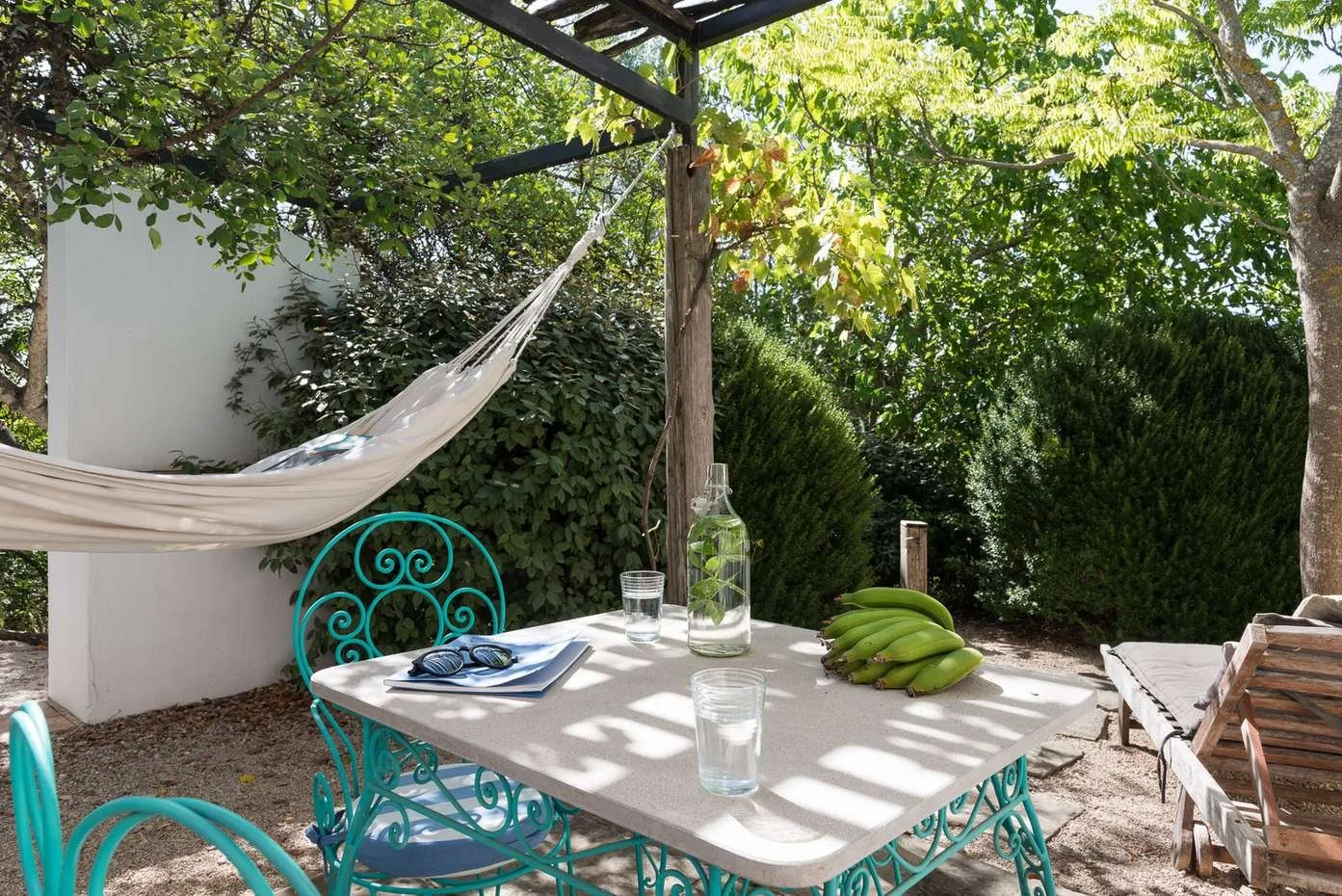 Outdoor patio scene with a white table, turquoise chairs, a glass bottle, glasses of water, bananas, a lounge chair, and a hammock, surrounded by green trees and bushes.