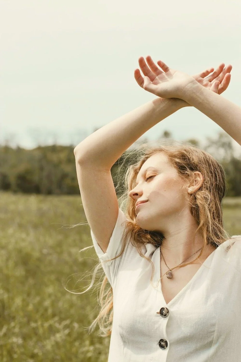 A young woman with red hair standing in a grassy field with her arms raised over her head, eyes closed, enjoying the sunlight.