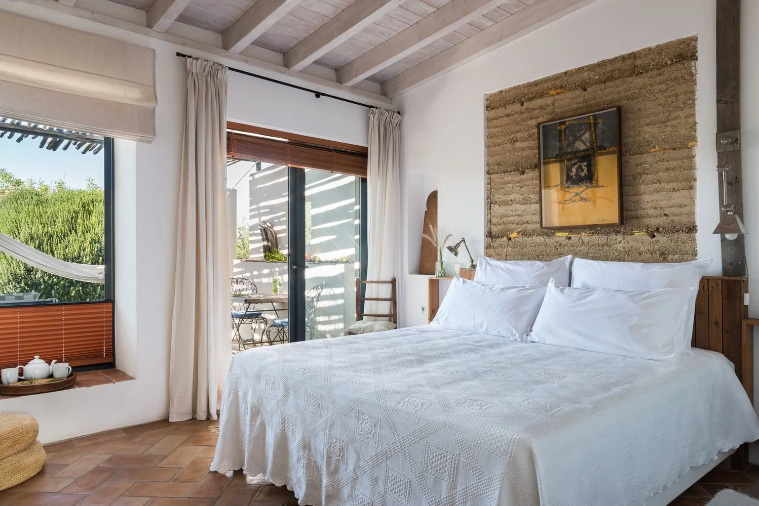 A cozy bedroom with a large white bed against a textured stone wall, natural light from sliding glass doors, and a small balcony outside with outdoor furniture.