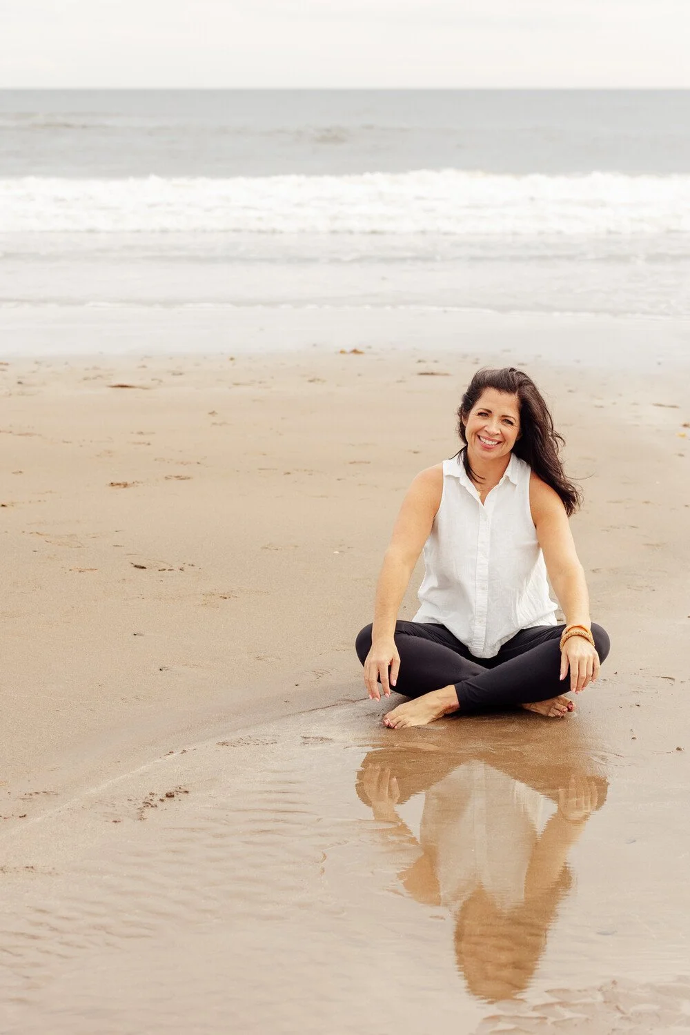 Woman sitting cross-legged on the wet sand at the beach, smiling, with ocean waves in the background.