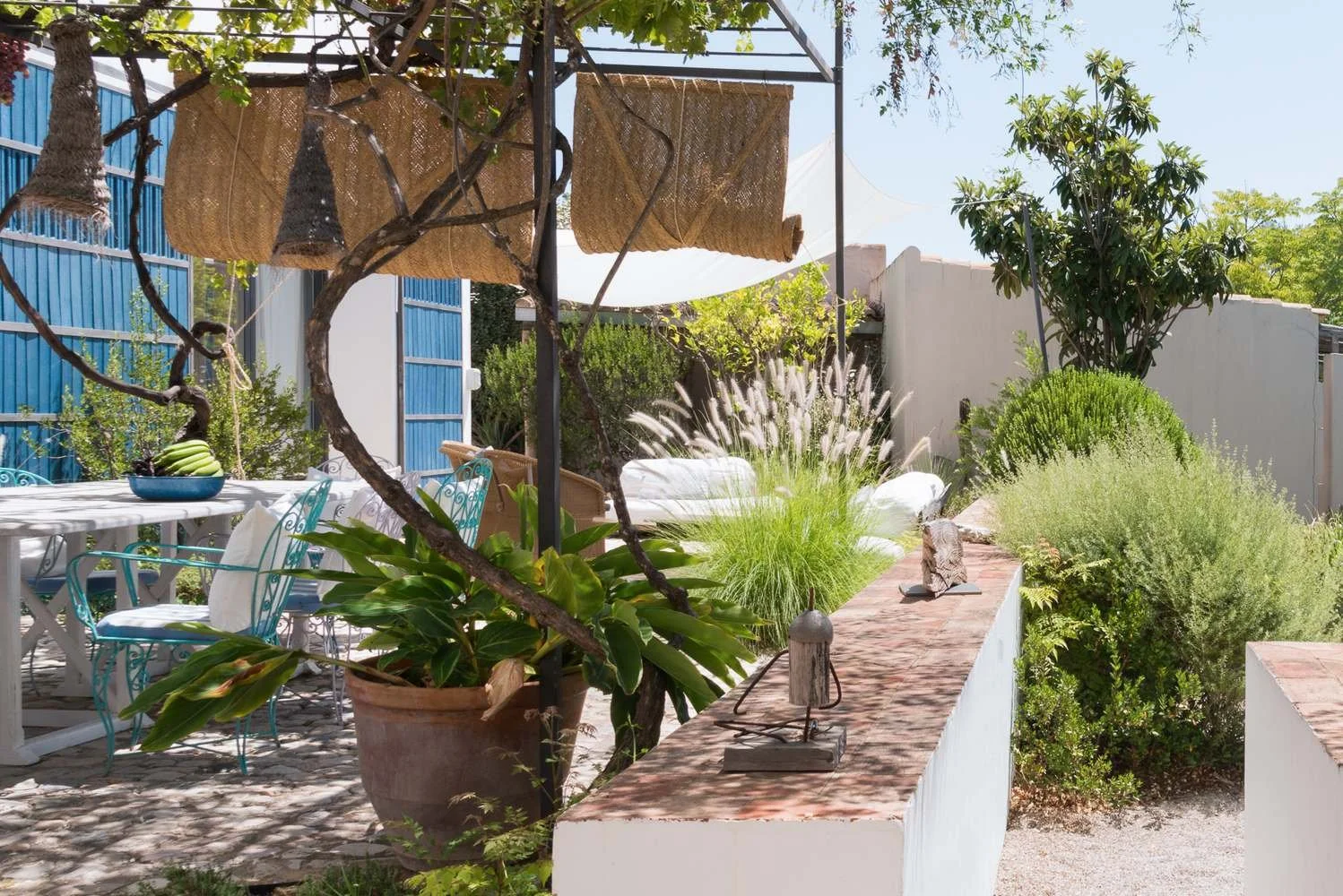 Outdoor patio with white dining table, turquoise chairs, potted plants, and lush greenery under sunlight.