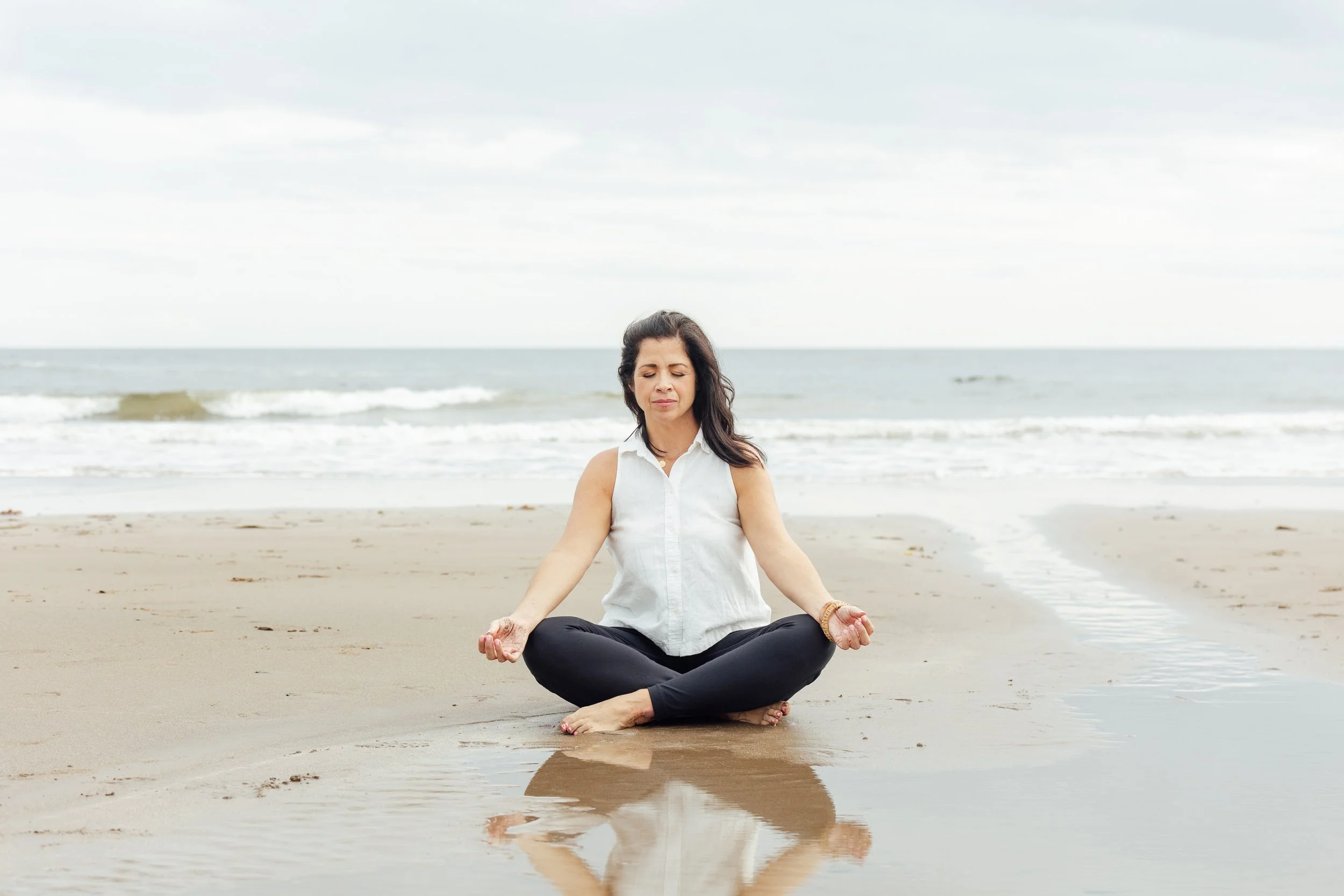 Charmaine Renee, rest coach and breathwork practitioner, practicing yoga on a sandy beach near the ocean, sitting cross-legged and meditating.