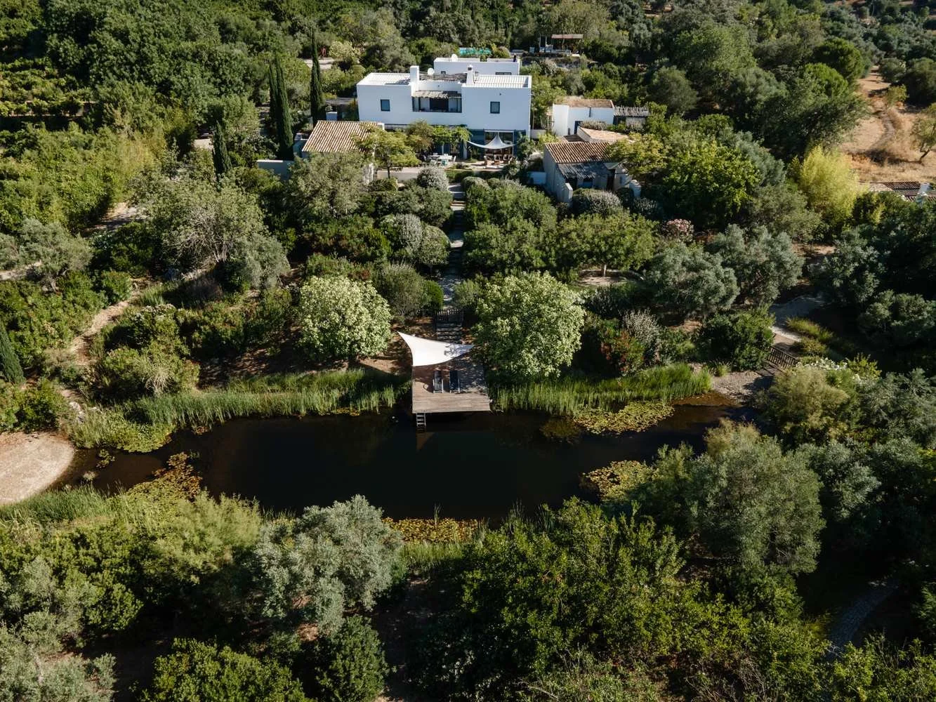 Aerial view of a modern white house nestled among dense trees, with a pond and wooden dock in the foreground.