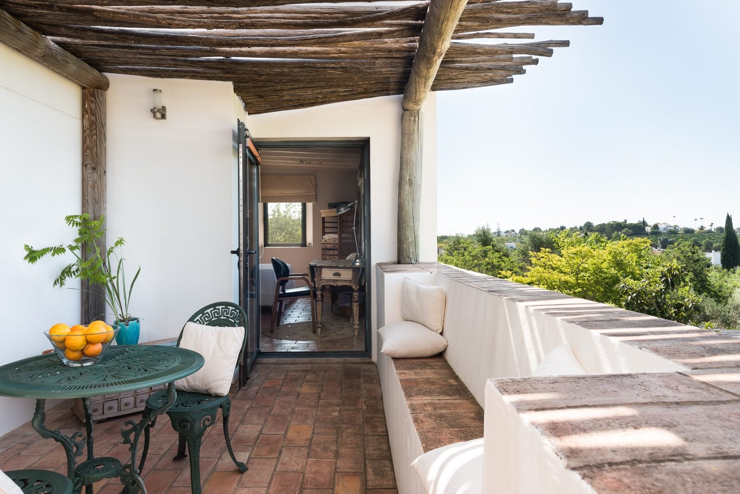 Outdoor balcony with white stucco walls, a green metal table and chairs, a bowl of oranges, and cushioned seating area. The balcony has a brick floor and a wooden pergola overhead. There's an open door leading inside a room with a desk and workspace.