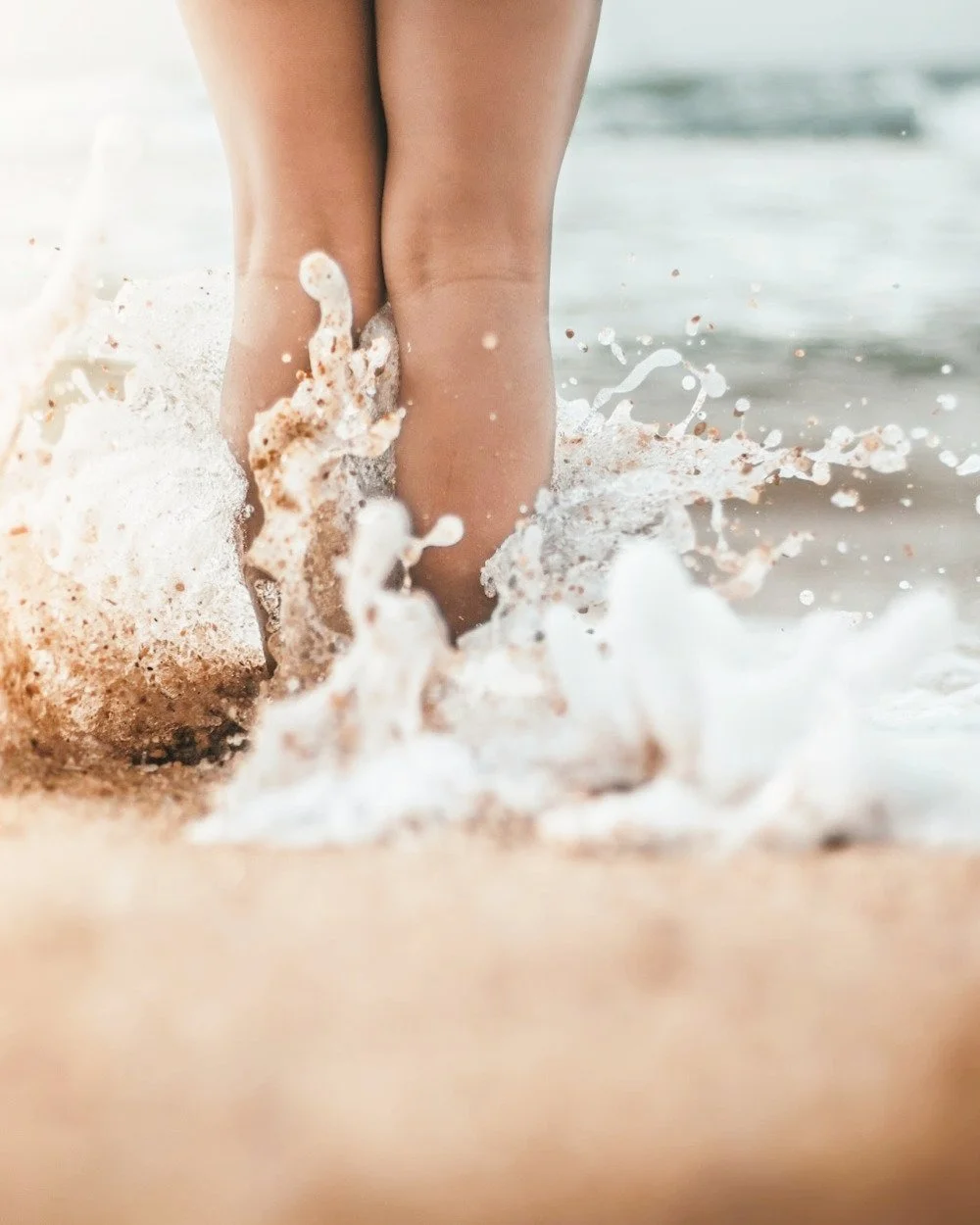 A person's legs in the water at the beach, with waves splashing around the ankles.