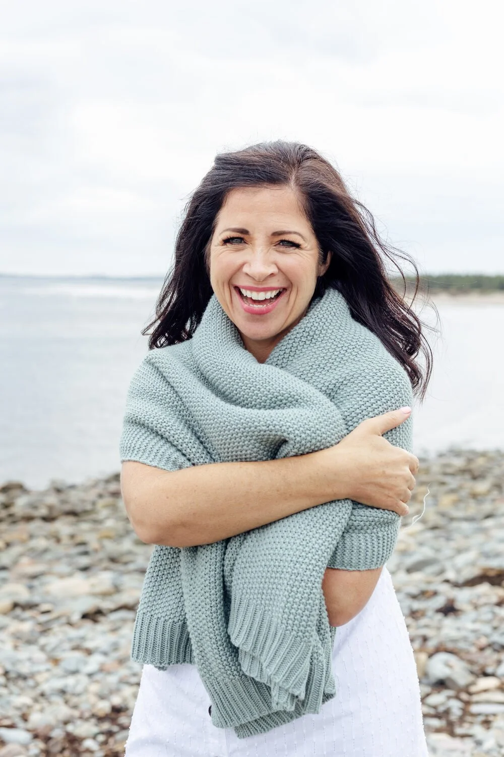 Charmaine Renee, rest coach and breathwork practitioner, smiling and hugging a gray knitted blanket while standing on a rocky beach with water and a cloudy sky in the background.