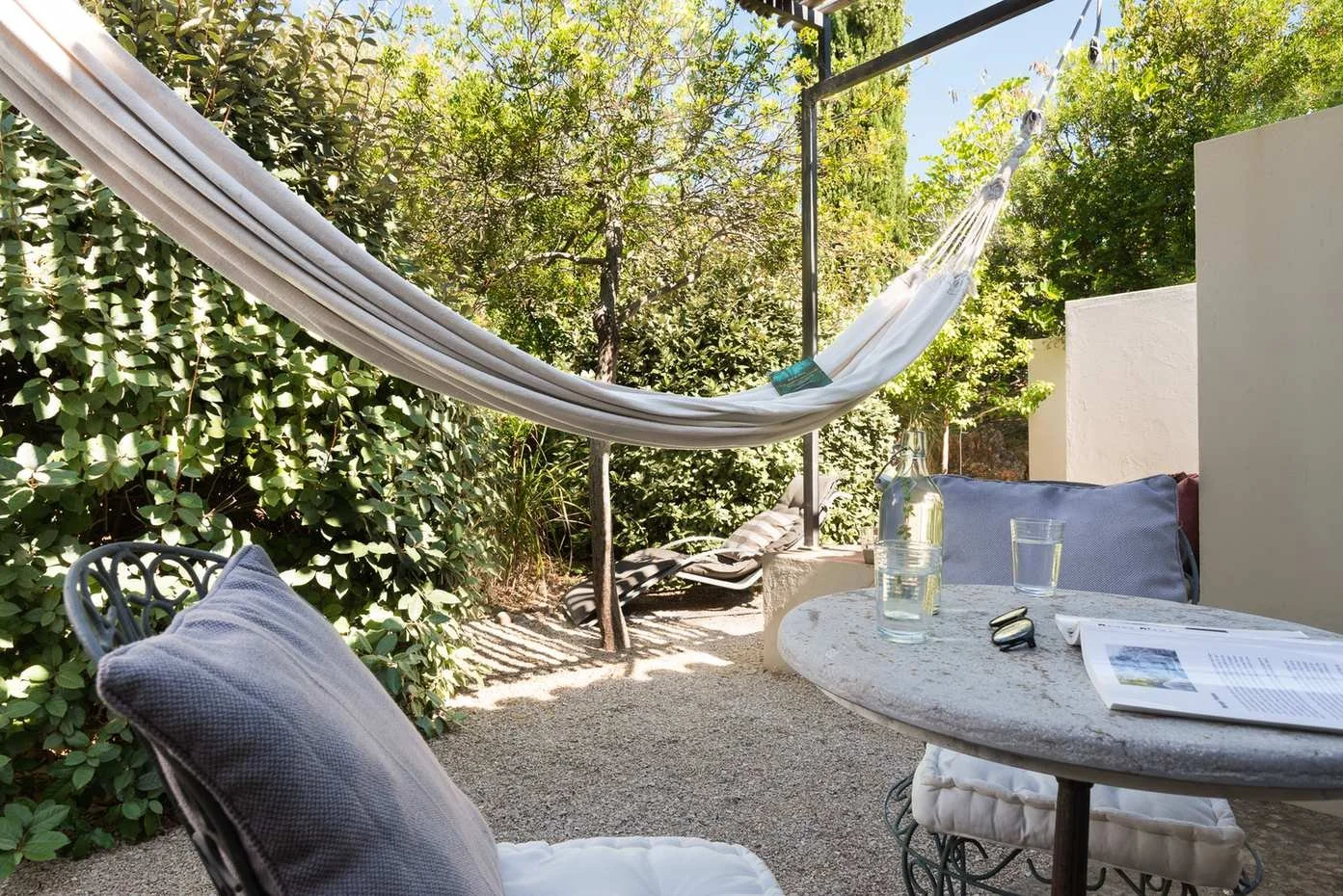 Outdoor patio with a hammock, cushioned chairs, a round table with a glass of water, a bottle, a newspaper, and a pair of sunglasses set against green bushes and trees.