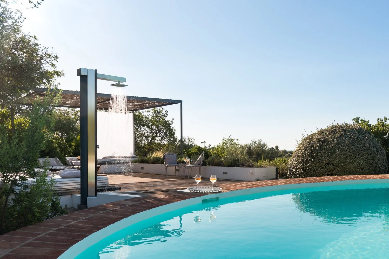 Outdoor pool with a brick edge, a modern outdoor shower, and a lounging area with chairs and cushions under a shaded structure, surrounded by greenery and clear sky.