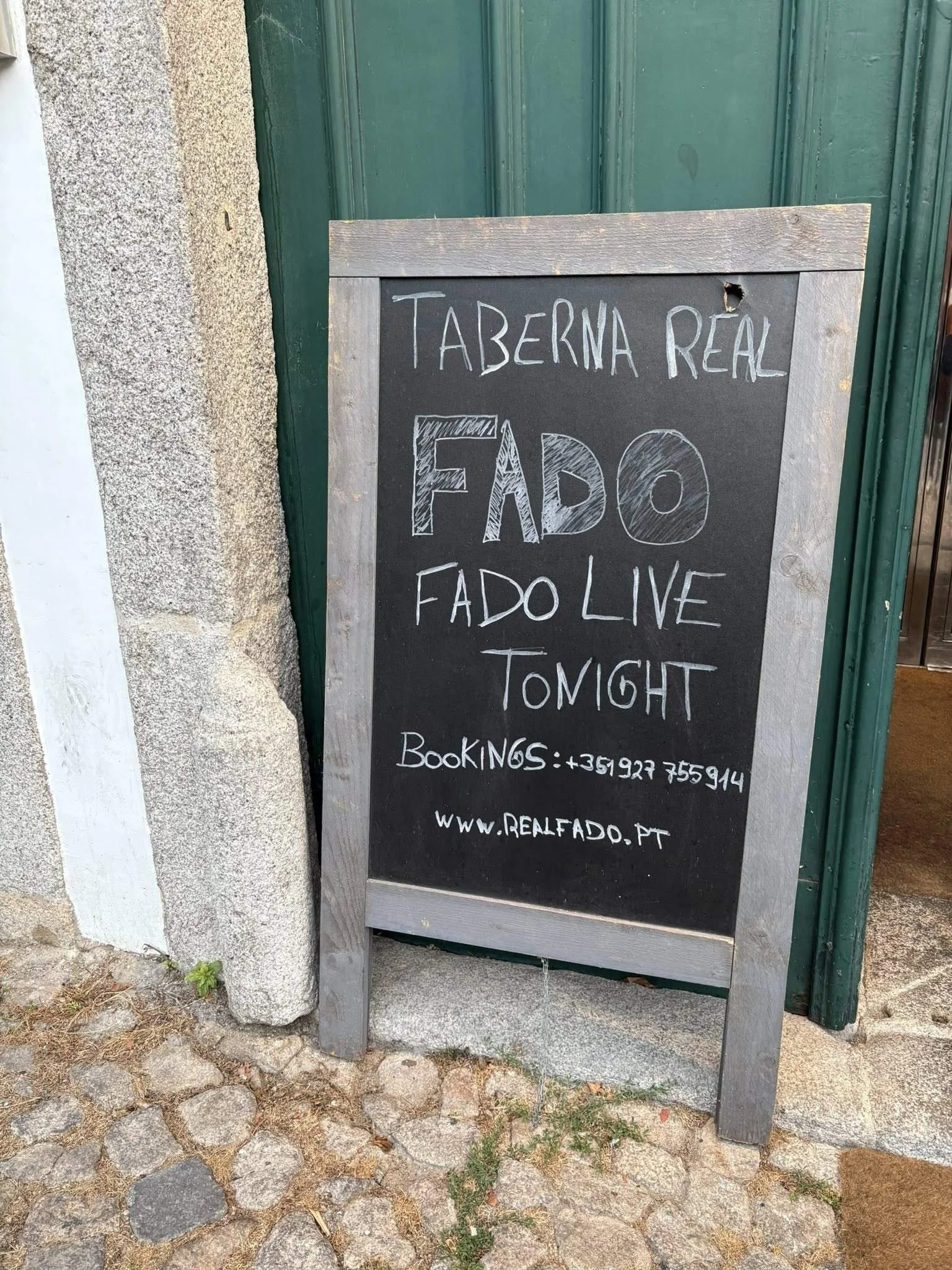 A chalkboard sign outside a restaurant titled 'Taberna Real' offering 'Fado Live Tonight' with contact number and website, placed on a cobblestone ground next to a stone wall and green door.