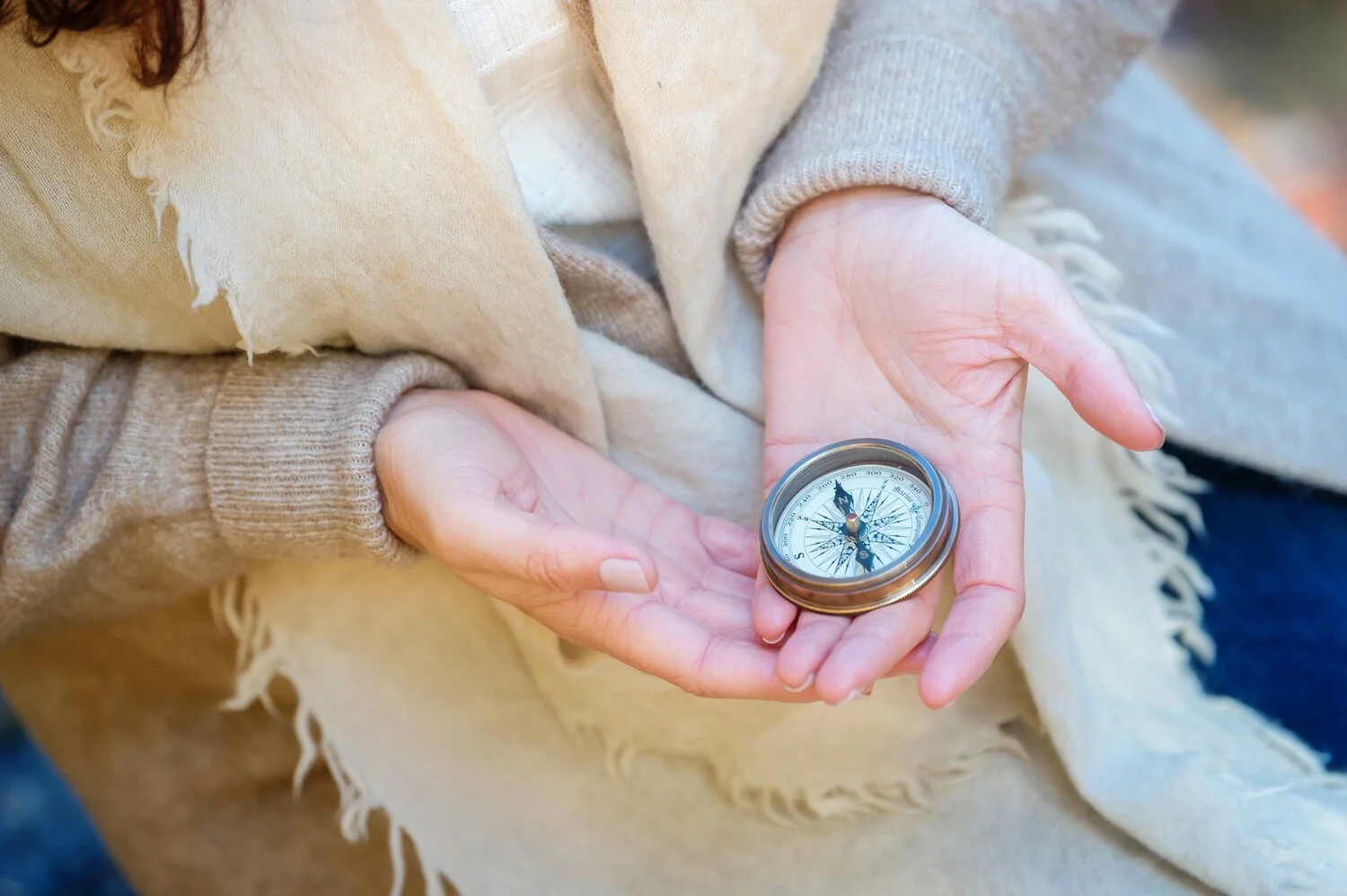 Person holding a compass in their hand, wearing a beige scarf with frayed edges and a gray sleeve.