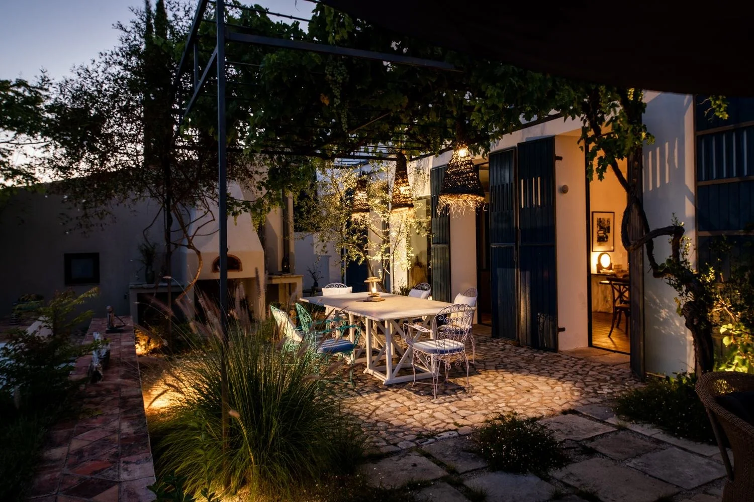 Outdoor patio with a dining table and chairs, lit by warm string lights and a lamp inside the house, surrounded by plants and a stone pathway during evening.