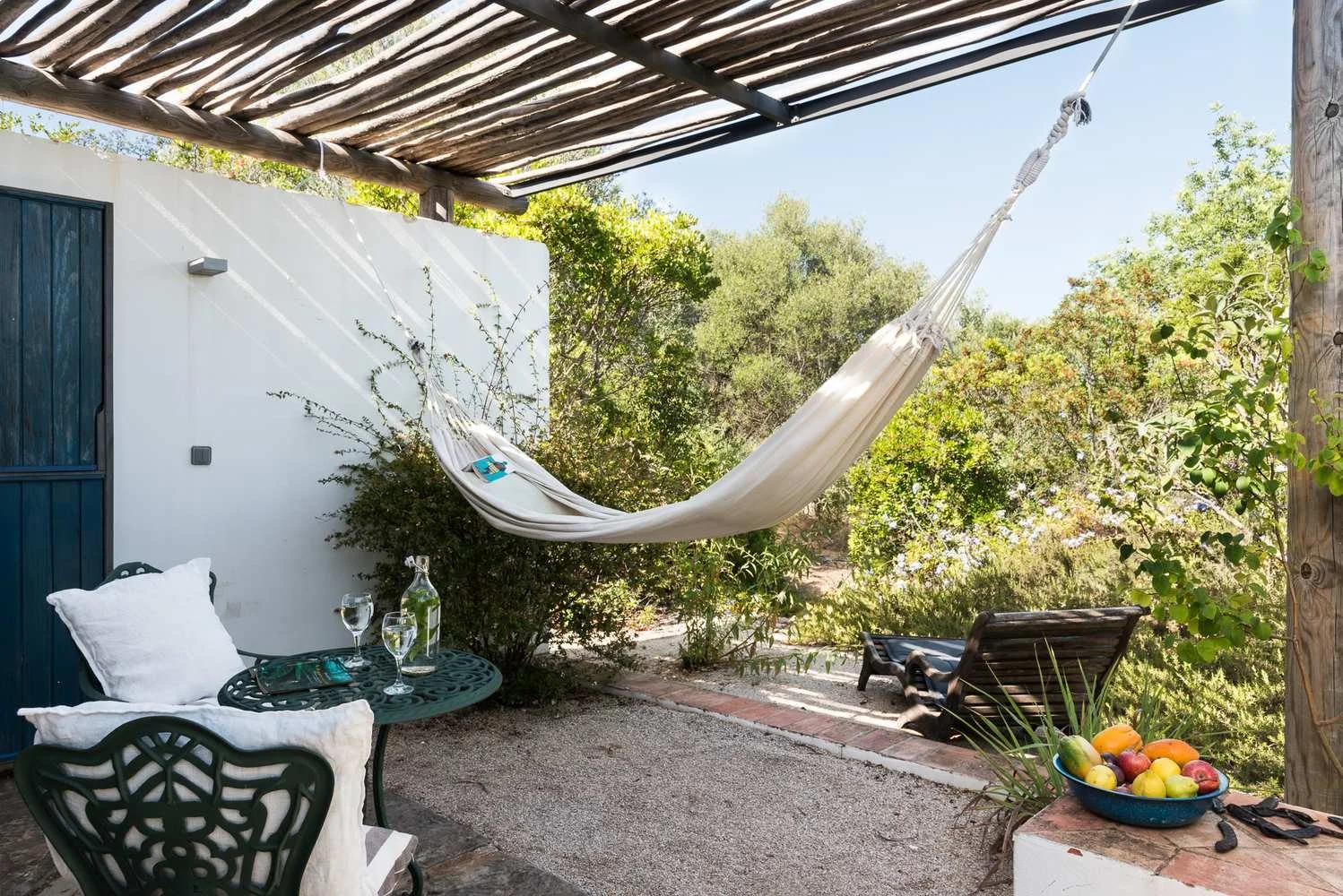 Outdoor patio with a white hammock, a black wrought iron table with two pillows, glasses, and a bottle of water. There is a bowl of fruit, a reclining chair, and green trees in the background.