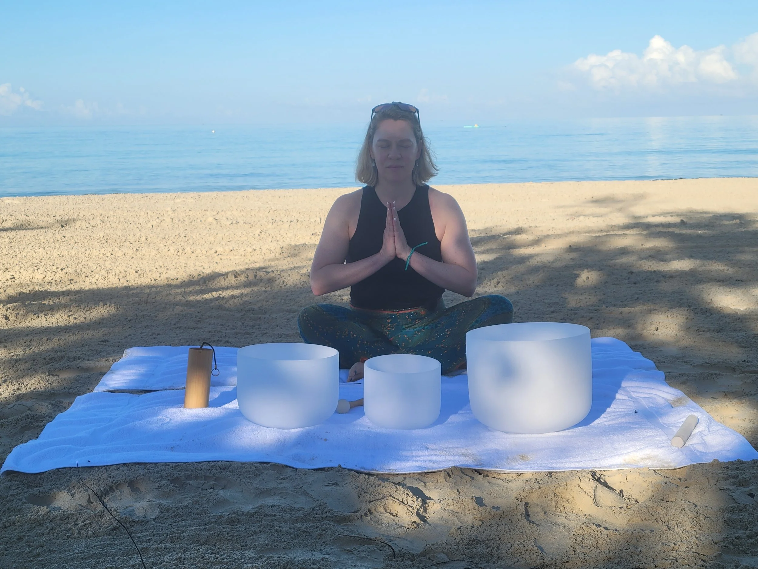 A woman with blonde hair and sunglasses on her head is sitting cross-legged on a blanket on the sandy beach, meditating with her hands in prayer position, surrounded by crystal singing bowls, with the ocean and blue sky in the background.