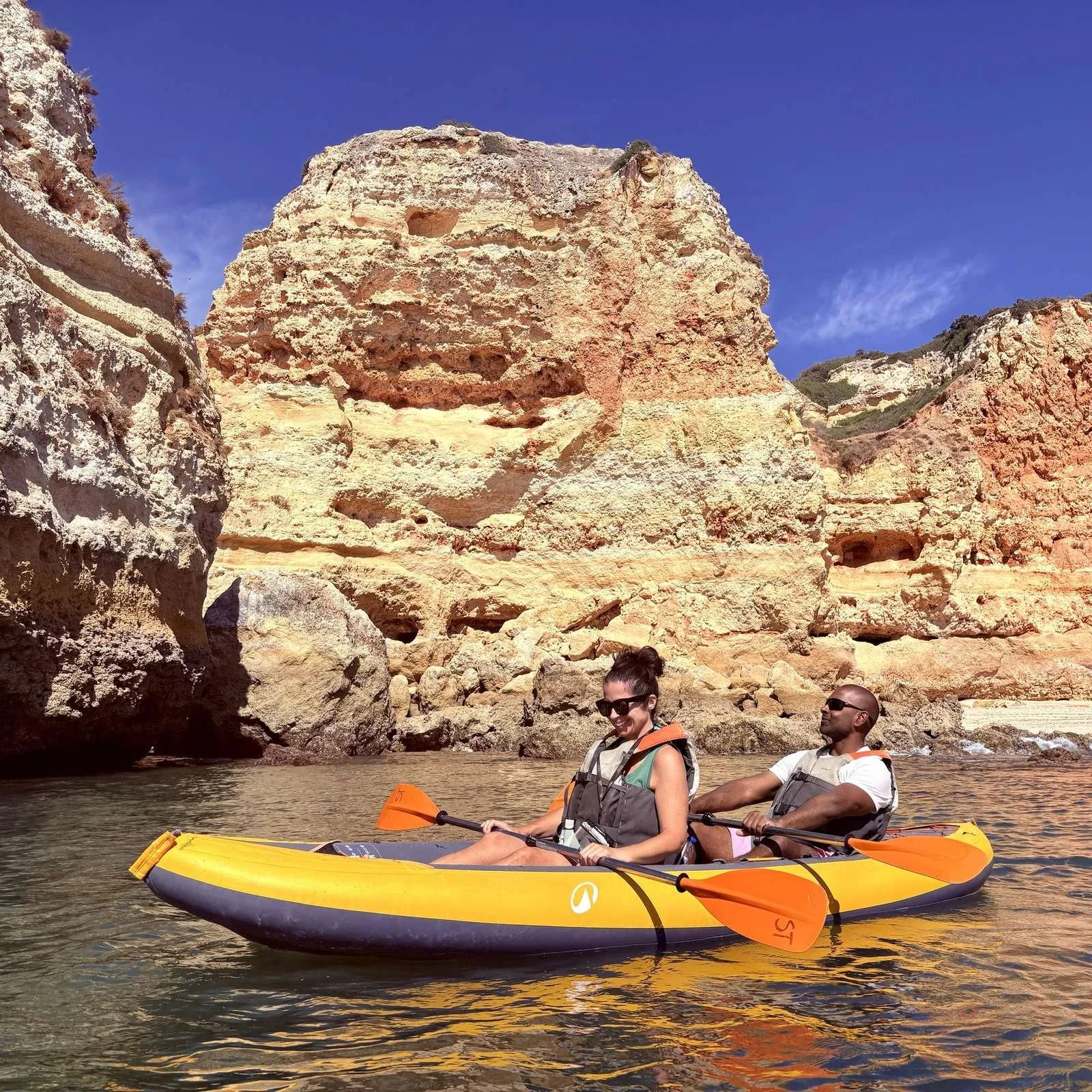 Two people kayaking on a river with tall rock formations and cliffs in the background under a clear blue sky.