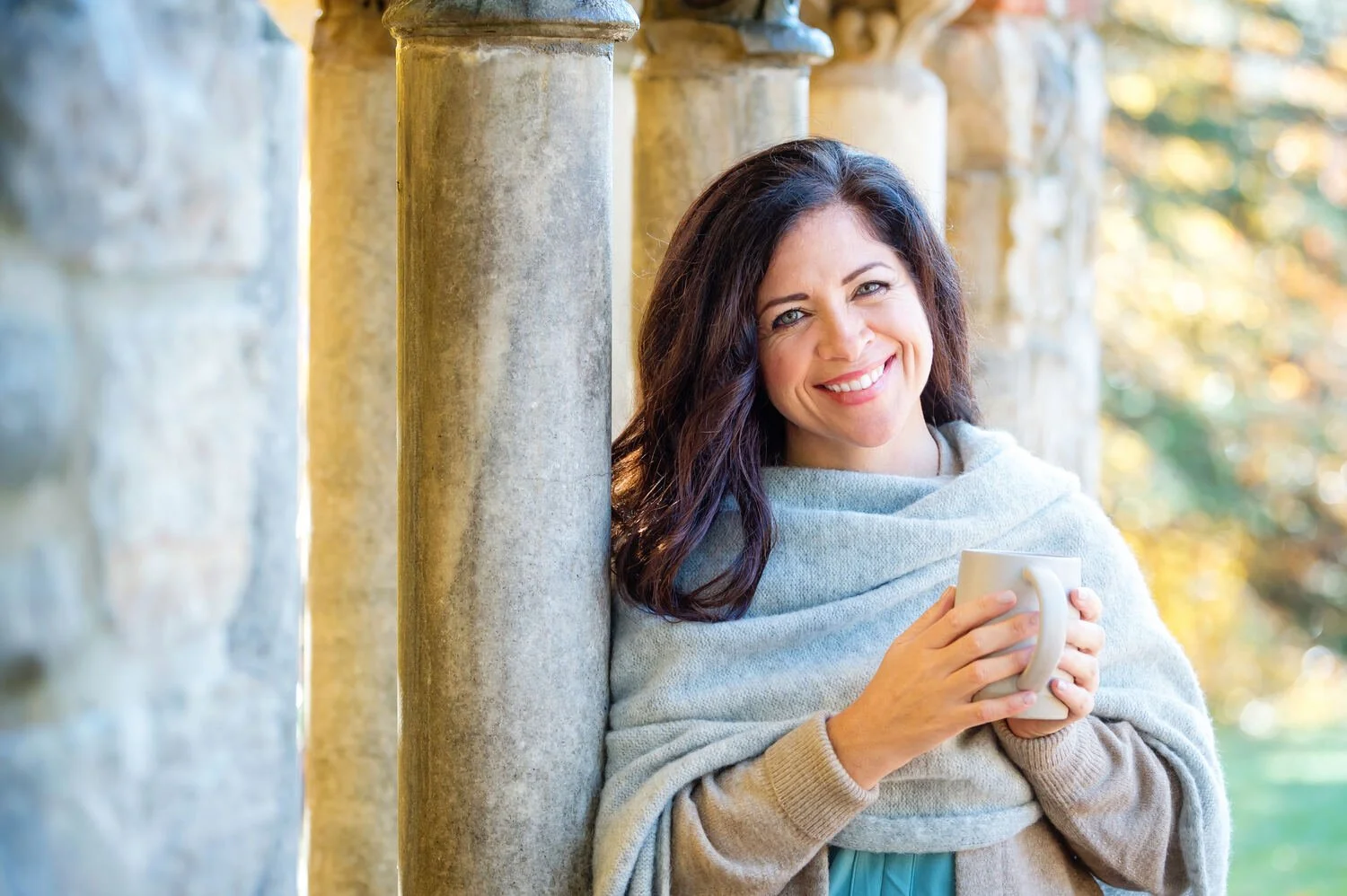 A woman with dark hair smiling and holding a mug, standing outdoors beside stone columns with trees in the background during fall.