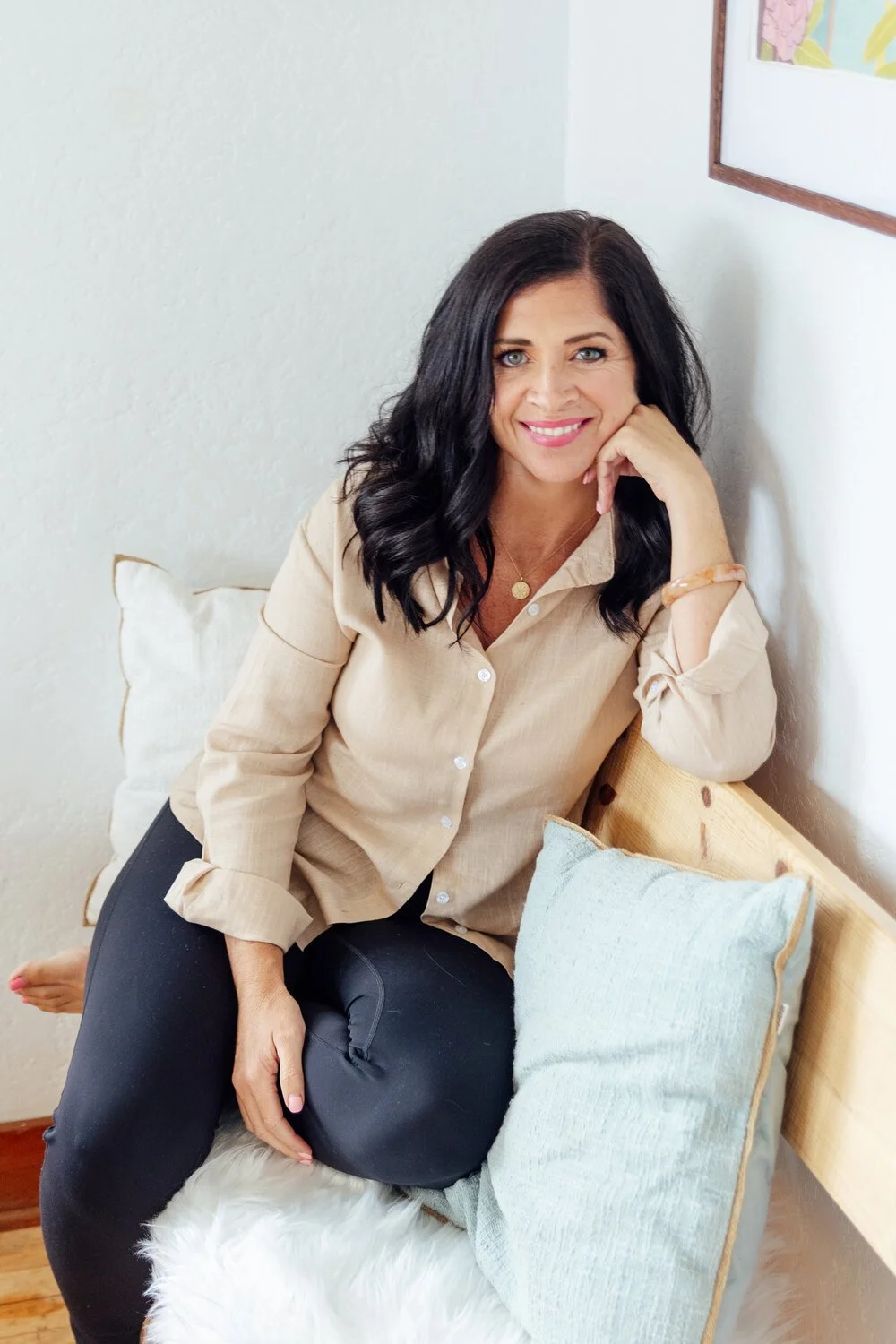 A woman with dark hair and blue eyes smiling while sitting on a cozy bench with cushions, dressed in a beige button-up shirt and black pants, in a well-lit room with framed artwork on the wall.