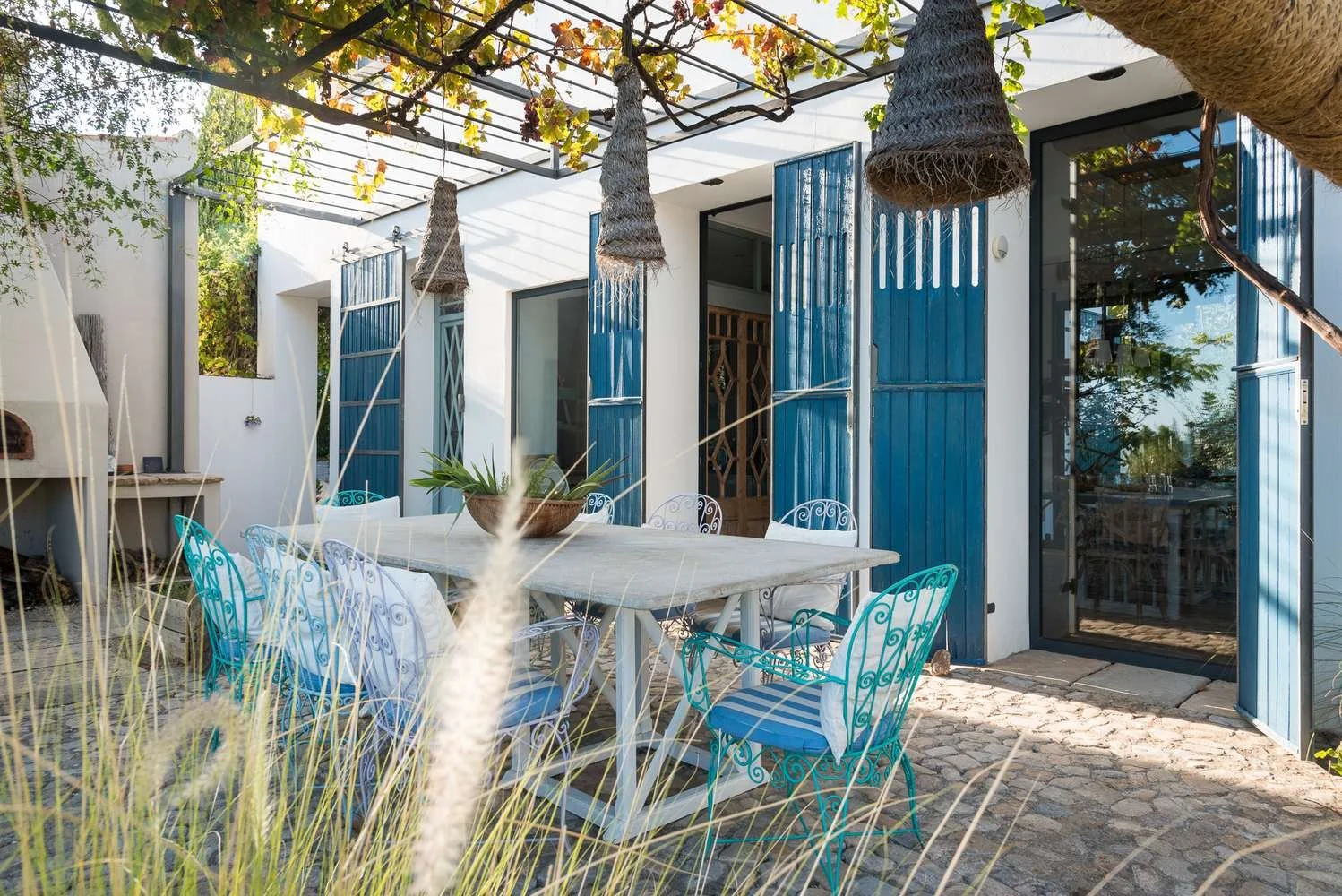 Outdoor patio area with a white table, turquoise and white chairs, potted plant centerpiece, and blue window shutters. There is a pergola overhead with climbing plants, and the ground is paved with cobblestones.