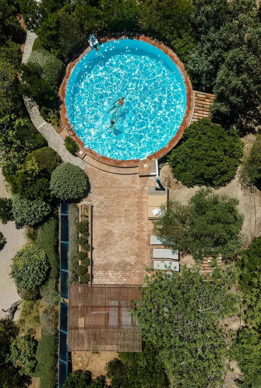A circular swimming pool with crystal-clear blue water, surrounded by greenery and trees, viewed from above.