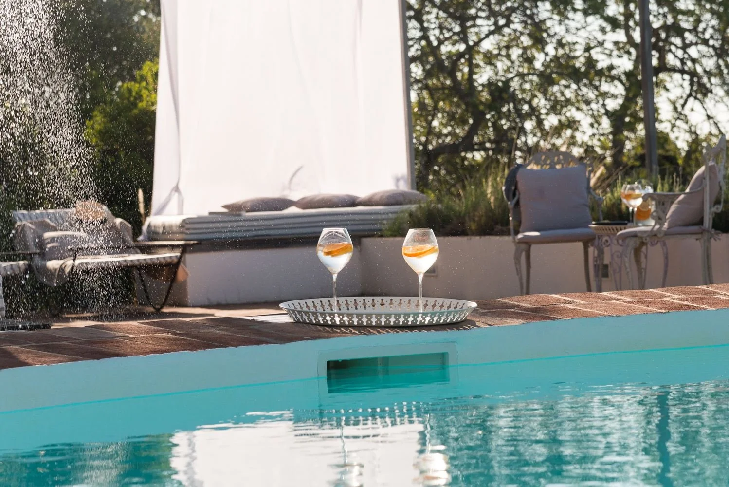 Two glasses of wine on a white tray near a swimming pool, with outdoor chairs and cushions in the background.