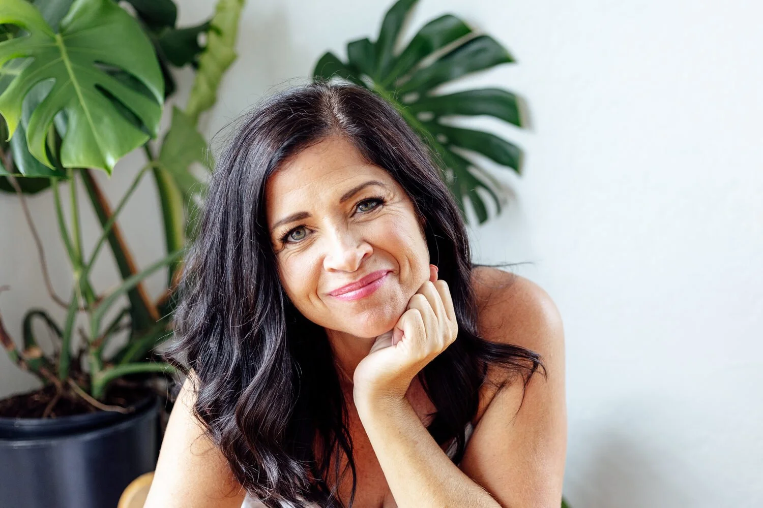 Charmaine Renee, rest coach and breathwork practitioner, smiling gently, resting her chin on her hand, with large green tropical plants in the background.