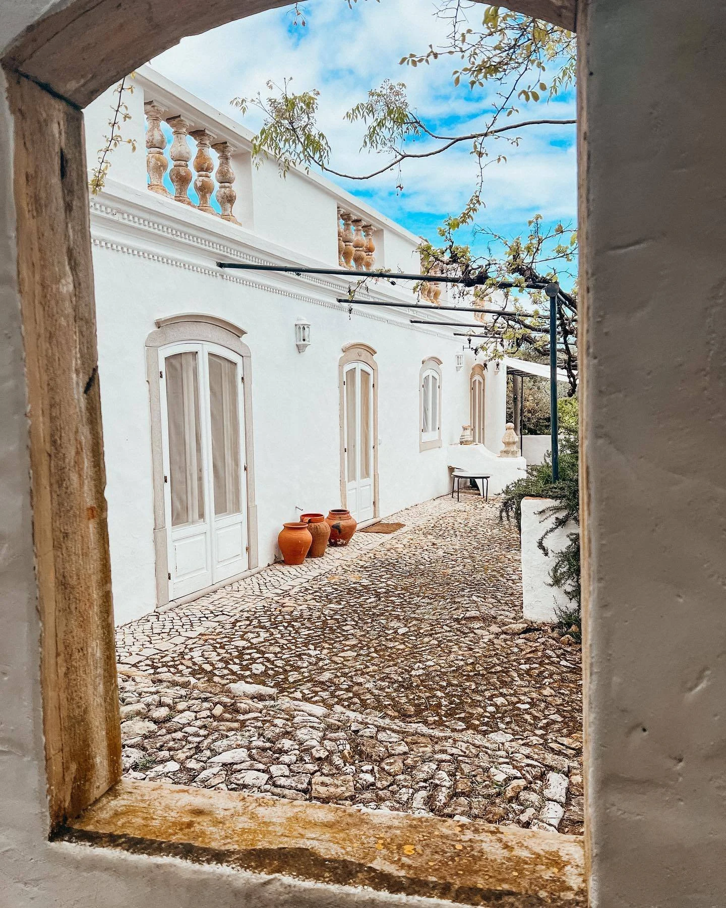 View of a white building with arched windows and terracotta pots on the ground, as seen through a stone-framed window or opening. The scene includes a cobblestone pathway and some tree branches with leaves against a blue sky with clouds.