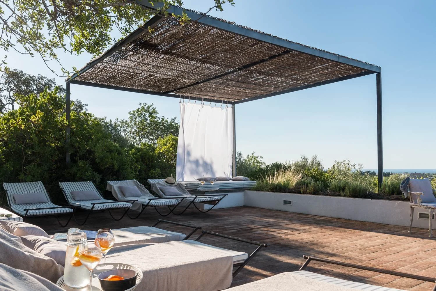Outdoor rooftop lounge area with white and black striped sun loungers, a white couch with drinks, and a canopy with hanging fabric for shade, surrounded by lush greenery and a view of the sea in the distance.