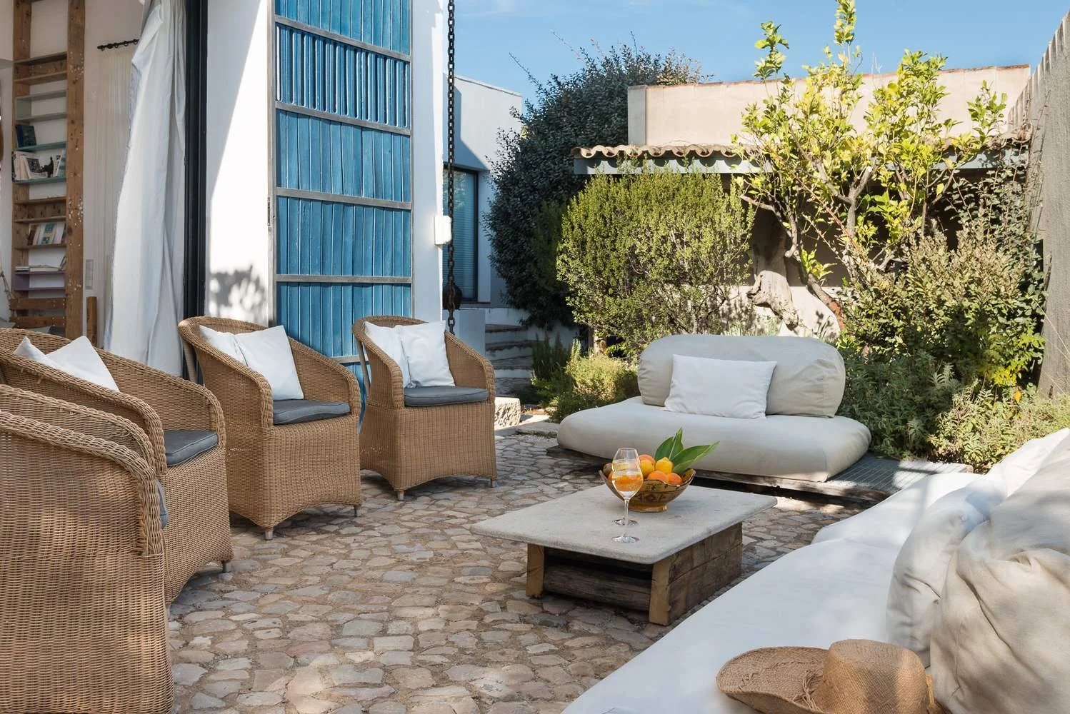 Outdoor patio with wicker chairs, a white sofa, and a small table with a fruit bowl and a glass of drink. There are plants and trees in the background, with a blue door on the left side.
