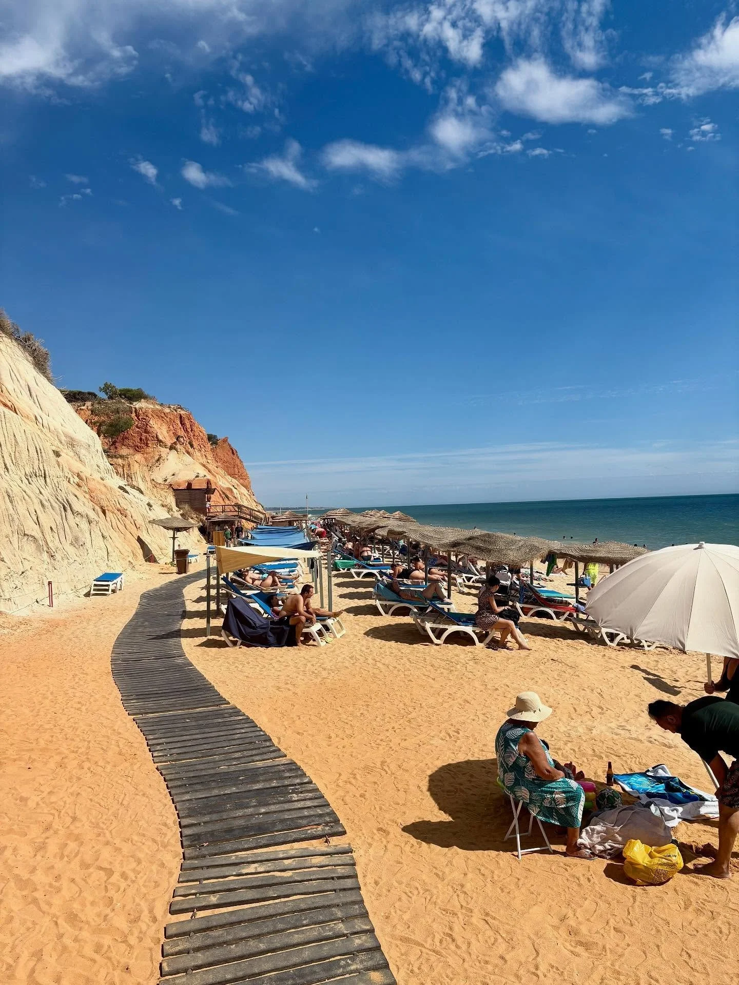 Beach scene with people relaxing on lounge chairs under umbrellas, a wooden pathway, sandy shore, and cliffs in the background under a partly cloudy blue sky.