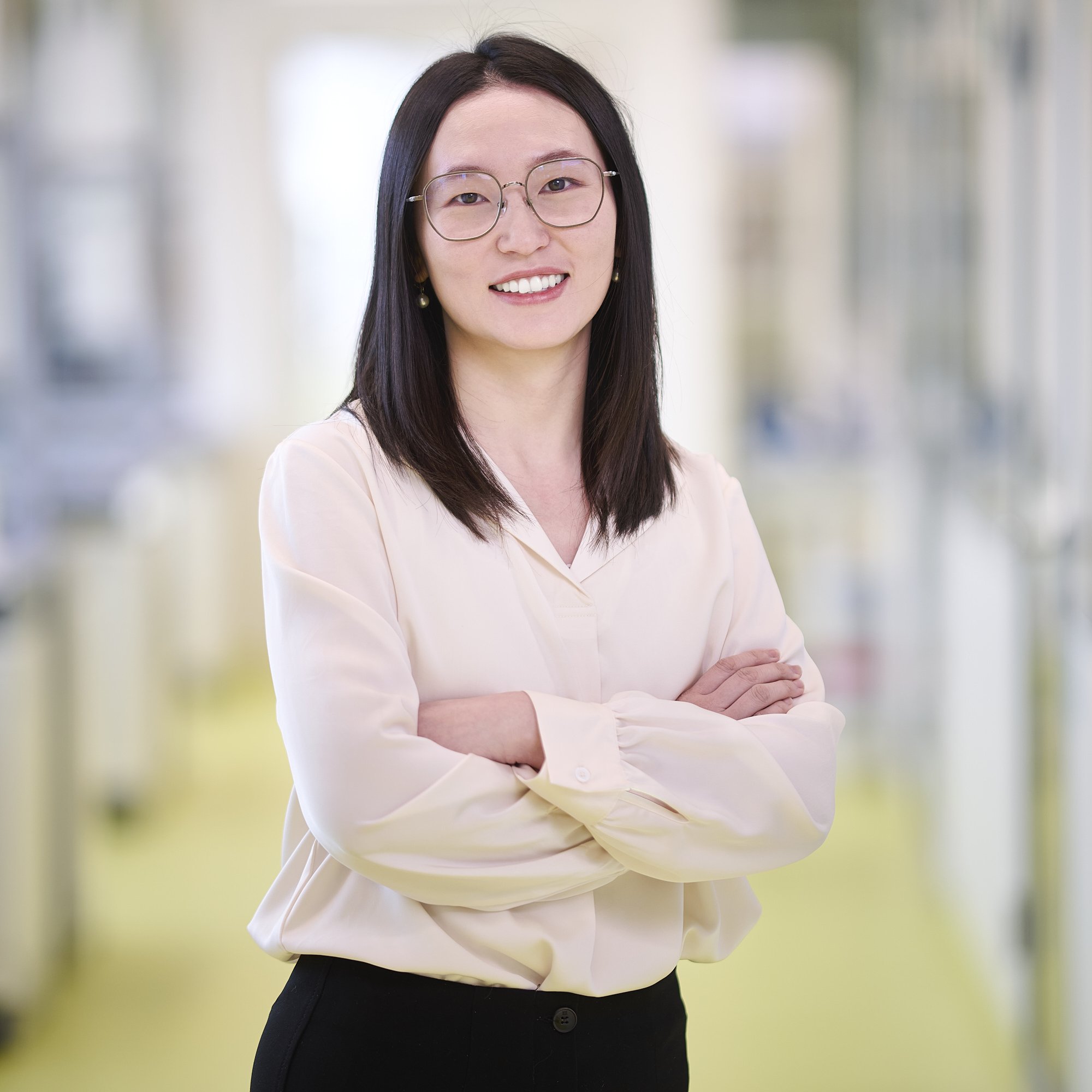 A woman with glasses and long dark hair smiling with arms crossed, standing in a well-lit modern office corridor.