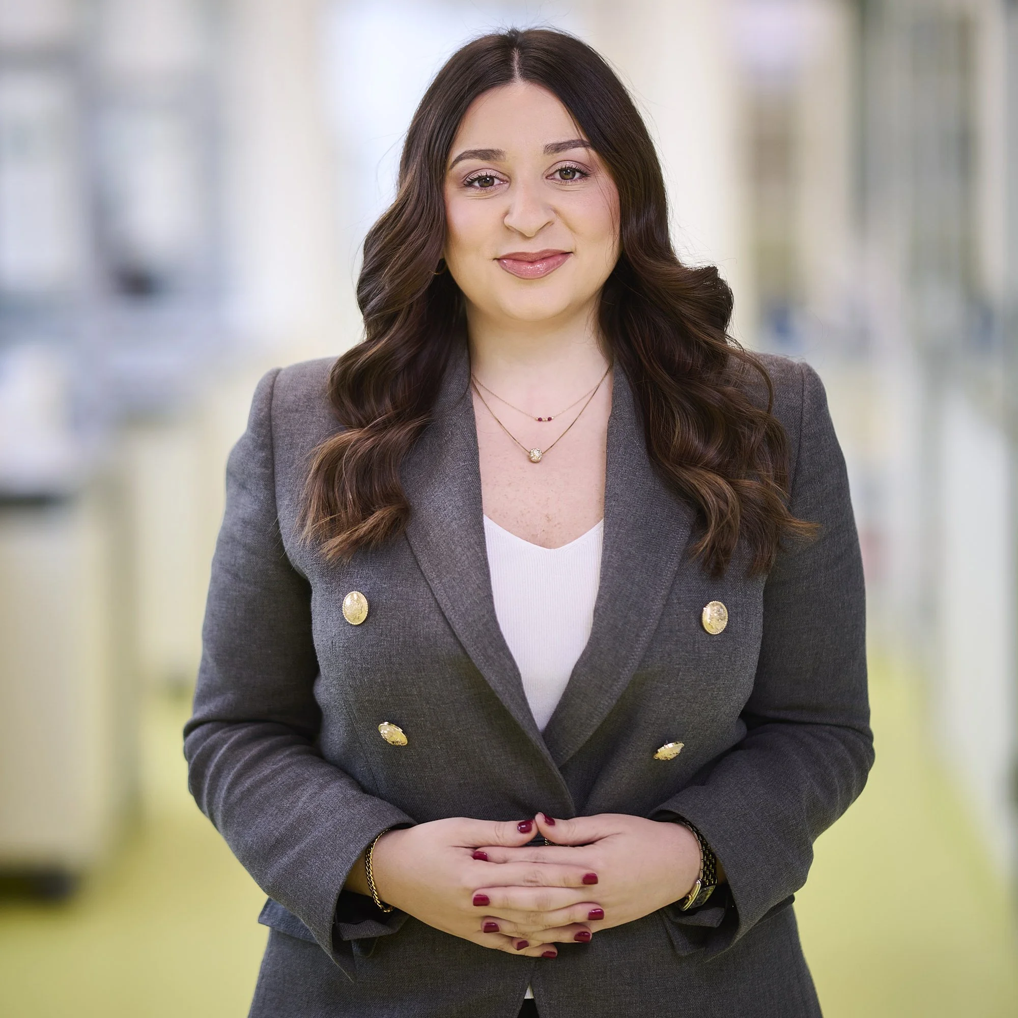 A woman in a gray blazer with gold buttons, wearing a white top, standing with her hands clasped in front of her, smiling at the camera in an indoor setting with blurred background.