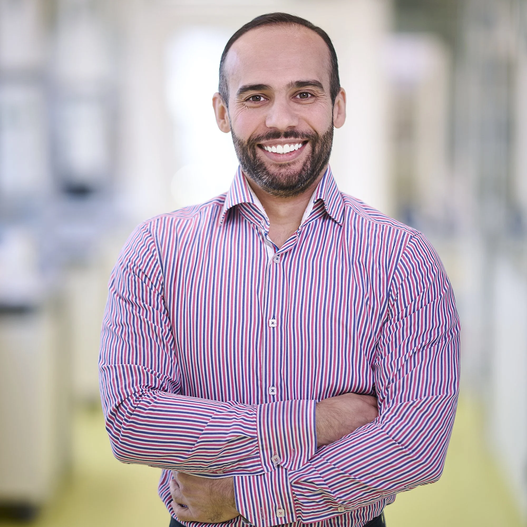 A smiling man with a beard wearing a striped button-up shirt, standing with arms crossed in an indoor setting with blurred background.